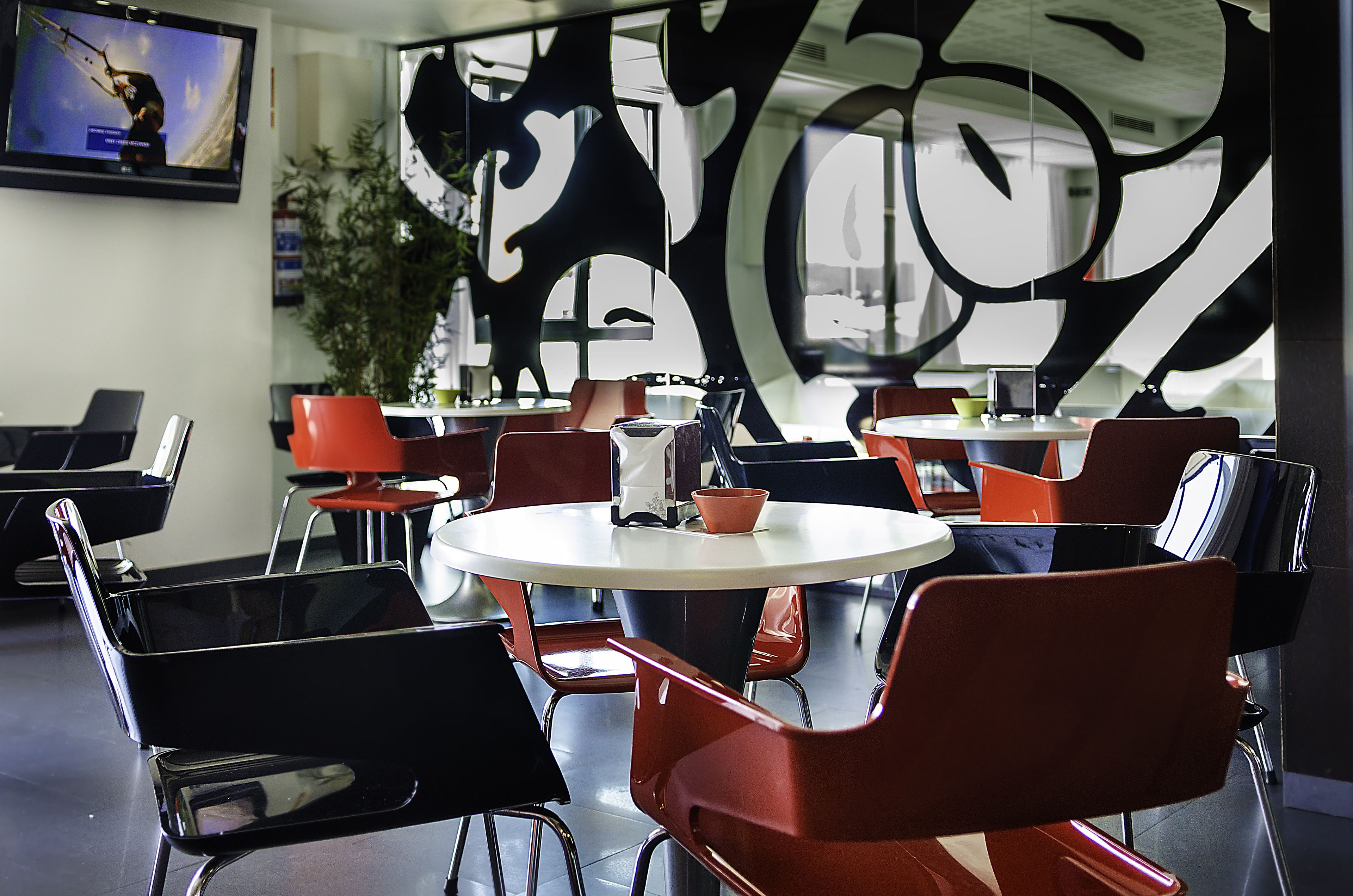 Un intérieur moderne avec des tables rondes et des chaises rouges, une grande œuvre d’art abstraite murale et un téléviseur fixé au mur.
