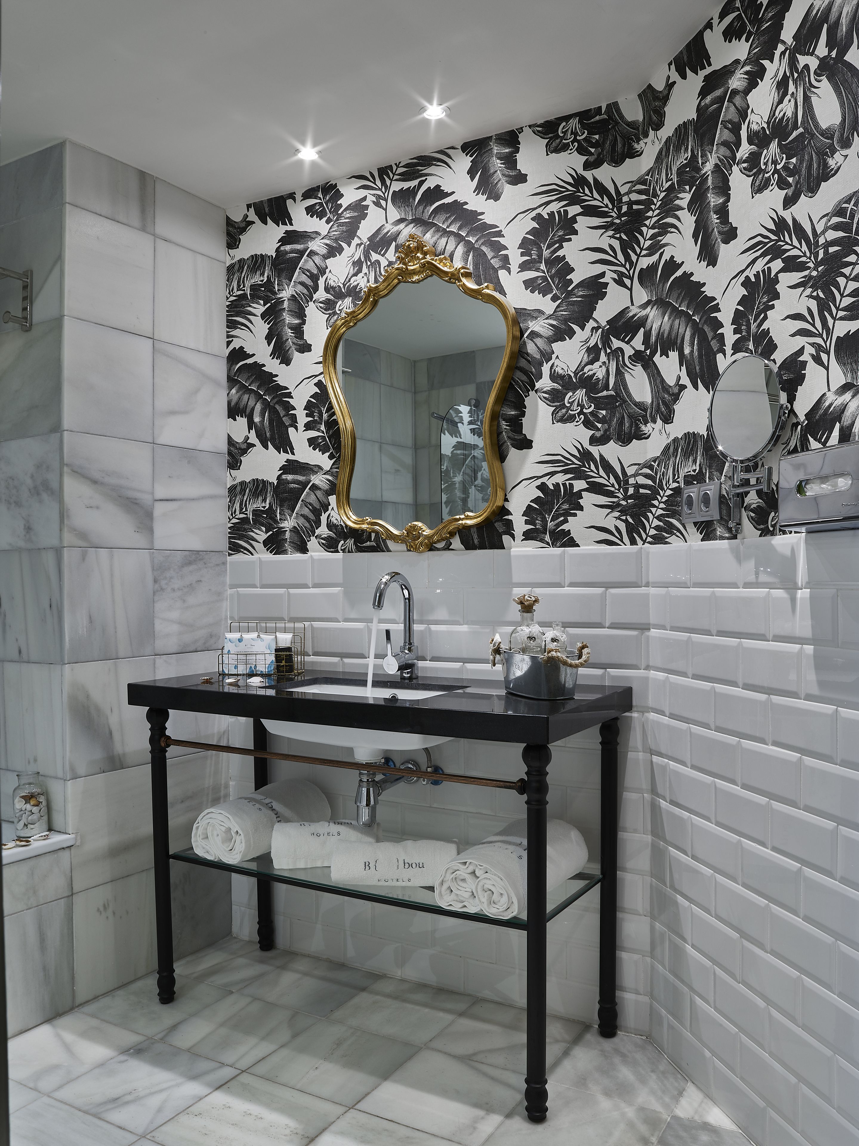 A bathroom with a black vanity, white marble tiles, a floral wallpaper, and a gold-framed mirror.