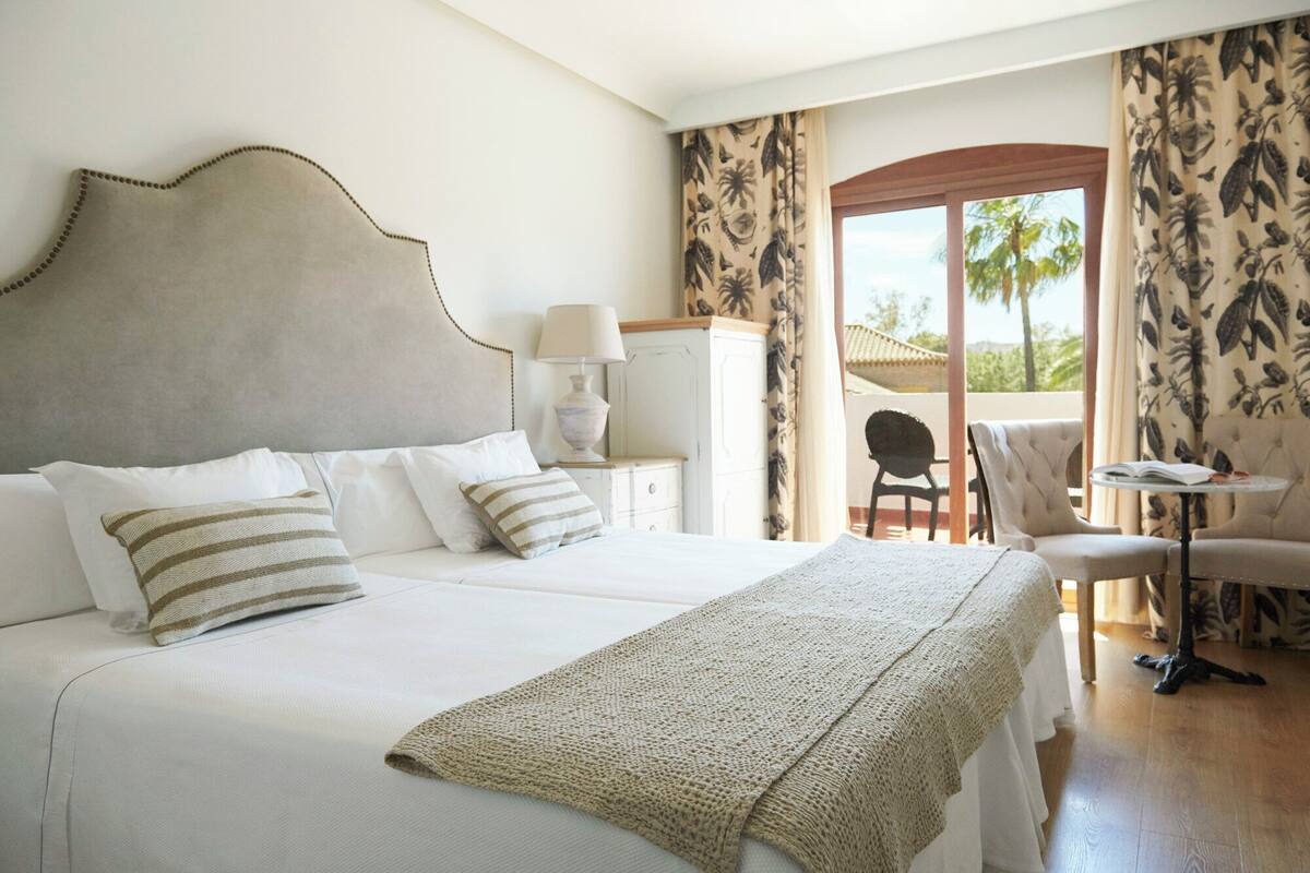 A hotel room with a large bed, a nightstand with a lamp, a chair, and a small table with a book.