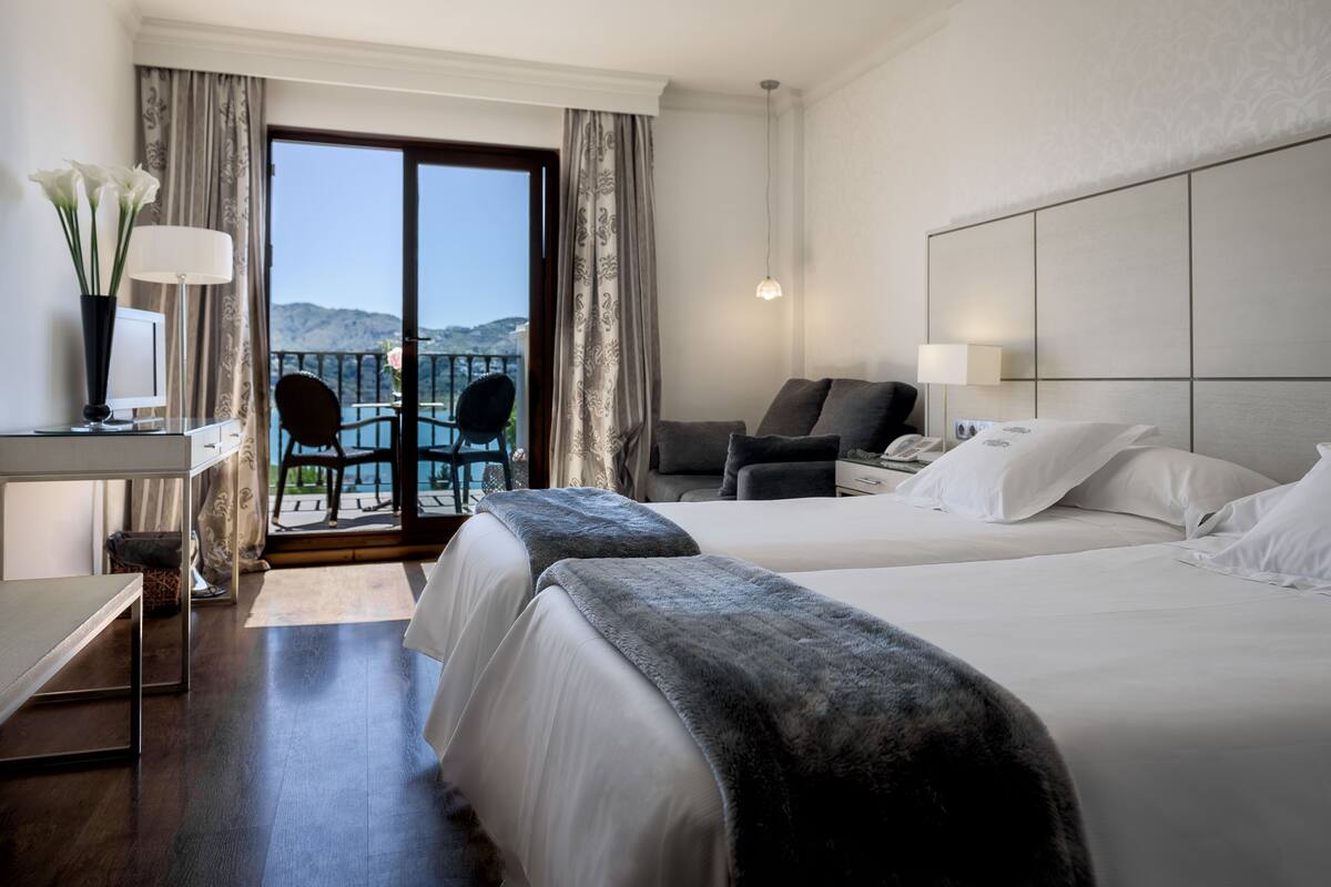 A hotel room with two beds, a desk with a computer, a chair, a balcony with a view, and a large mirror.