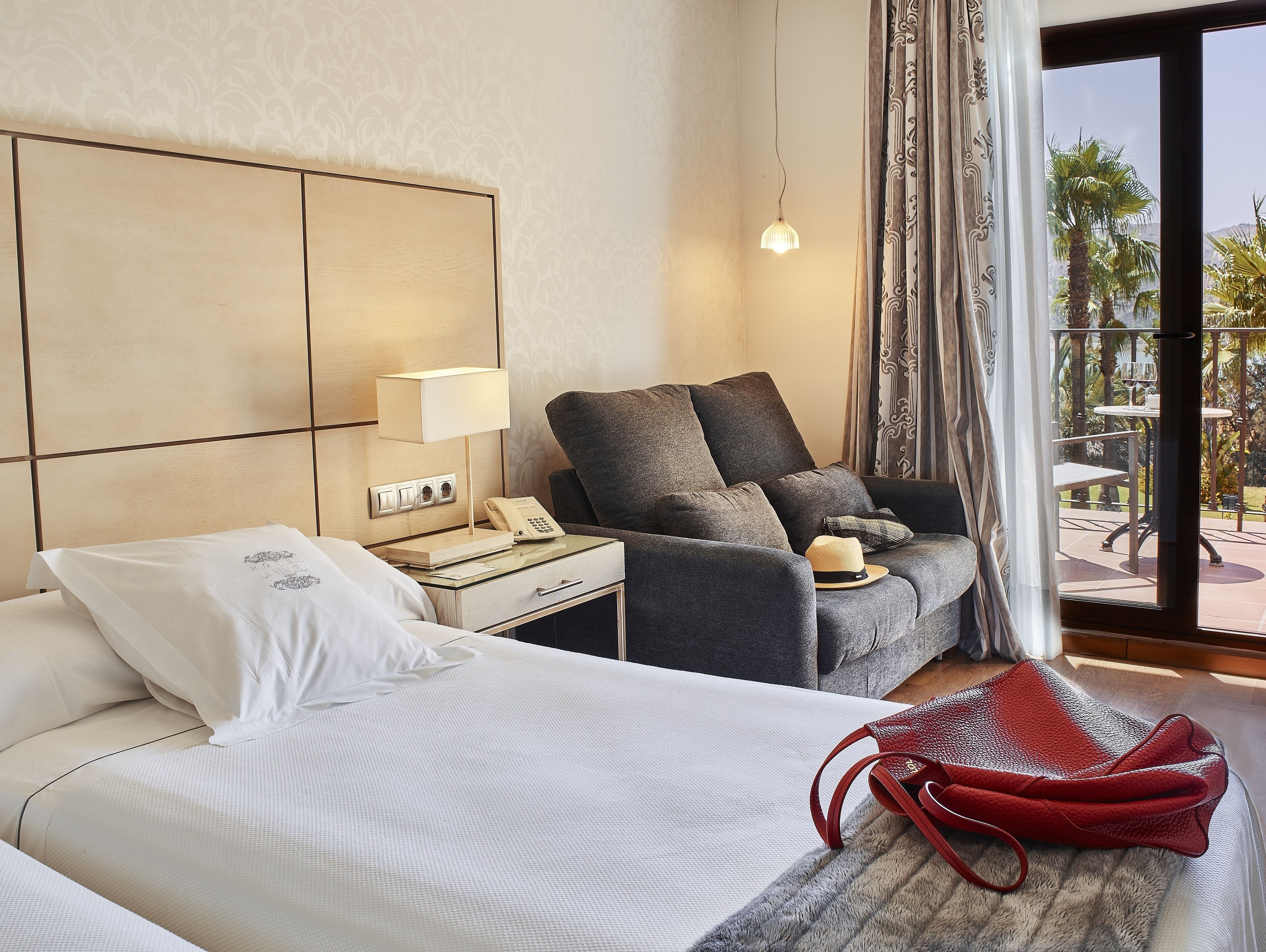 Ein Hotelzimmer mit Bett, Nachttisch mit Lampe, Stuhl und Blick ins Freie.