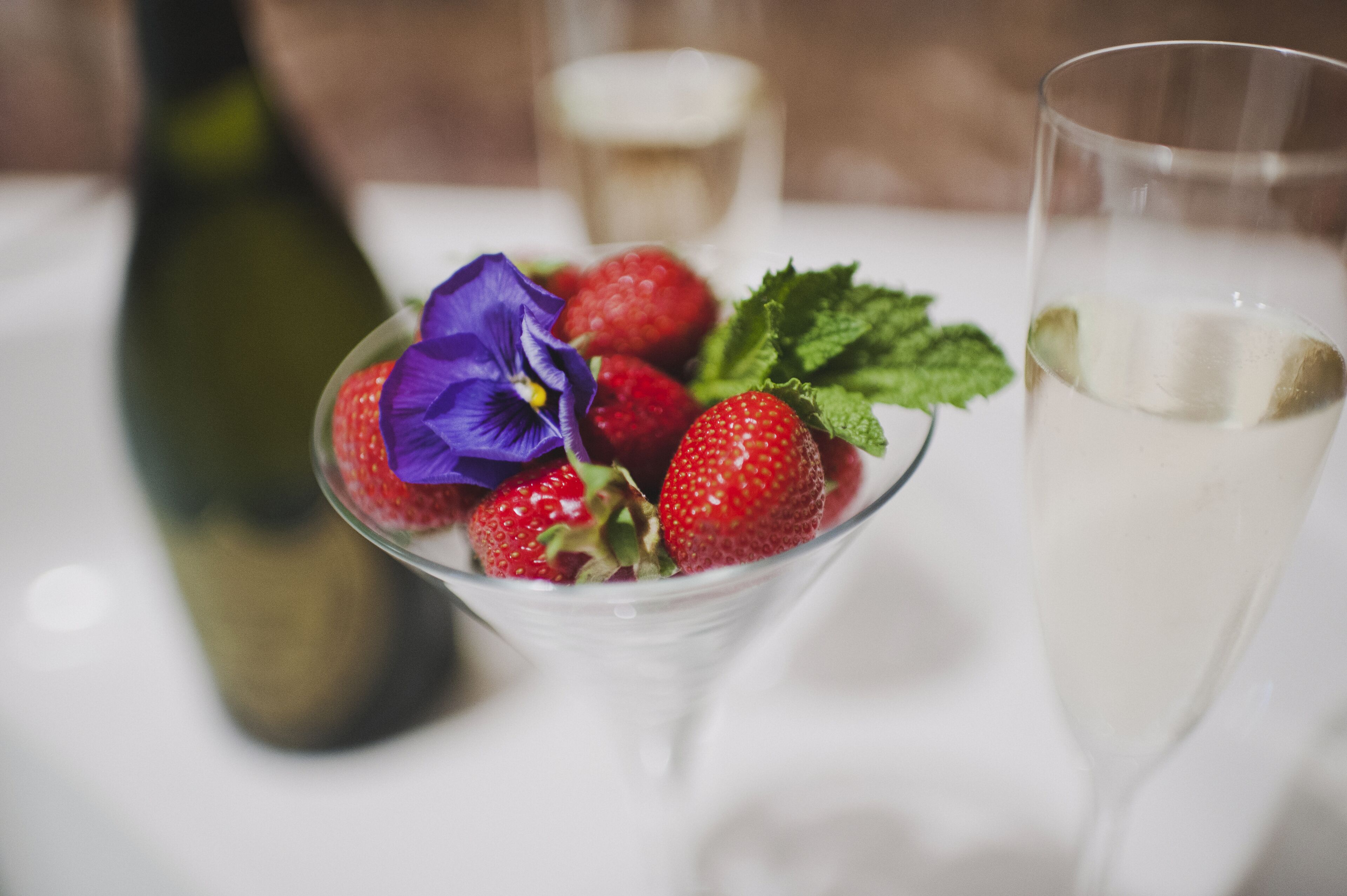 Um copo de martini com morangos e uma flor roxa, uma garrafa e um copo com líquido branco.