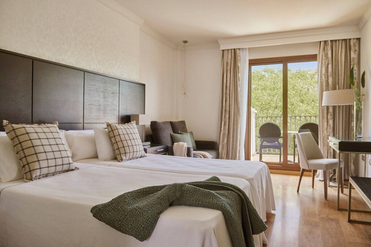 A hotel room with a large bed, a desk, a chair, and a balcony with a view of greenery.