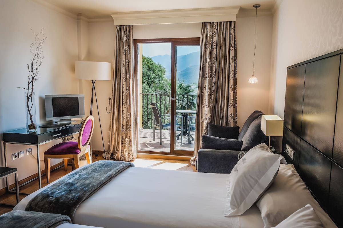 A hotel room with a bed, desk, chair, TV, and a balcony with a table and chairs.
