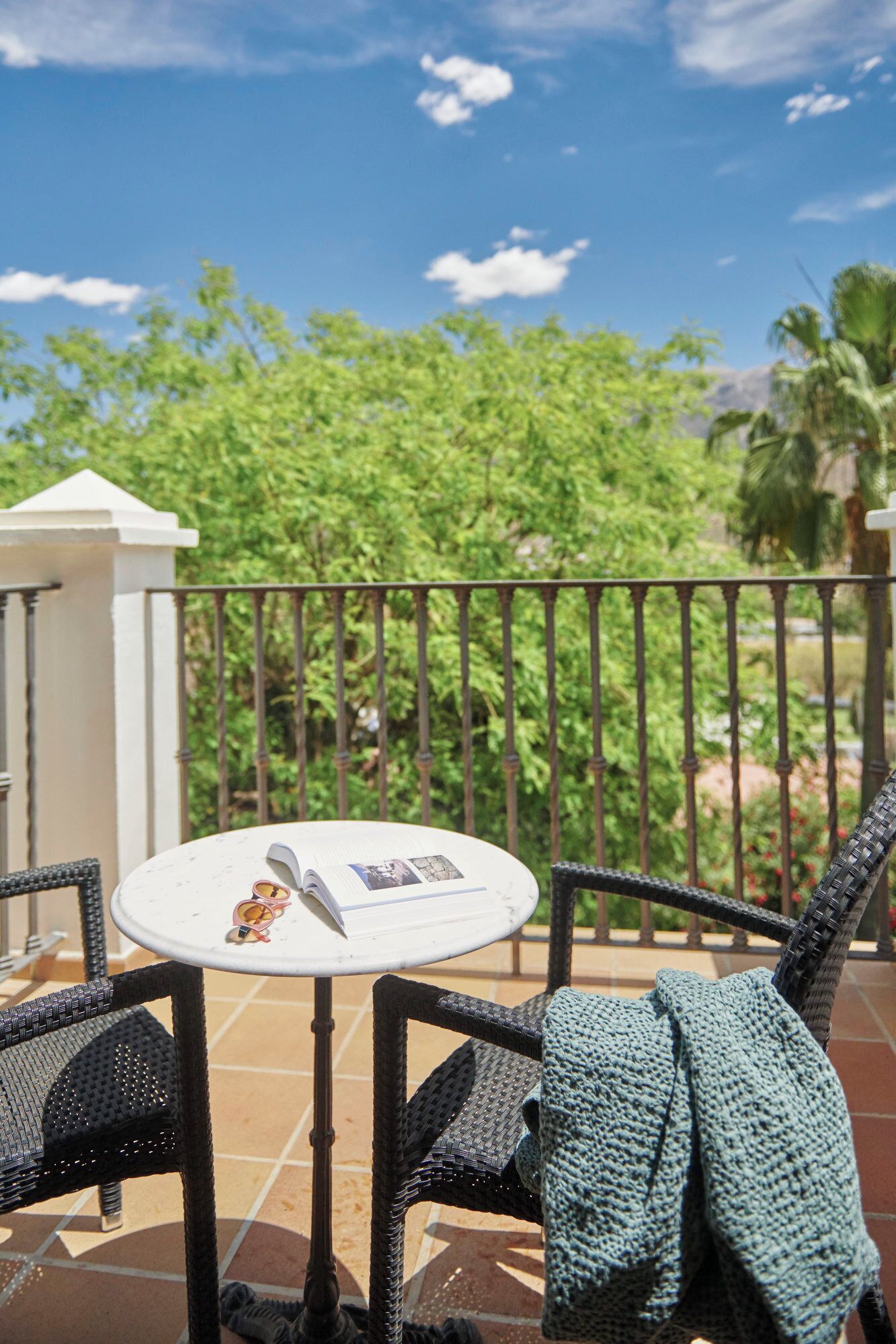 A round table with a book and sunglasses on it, surrounded by wicker chairs, on a balcony with a view of green trees and a clear sky.