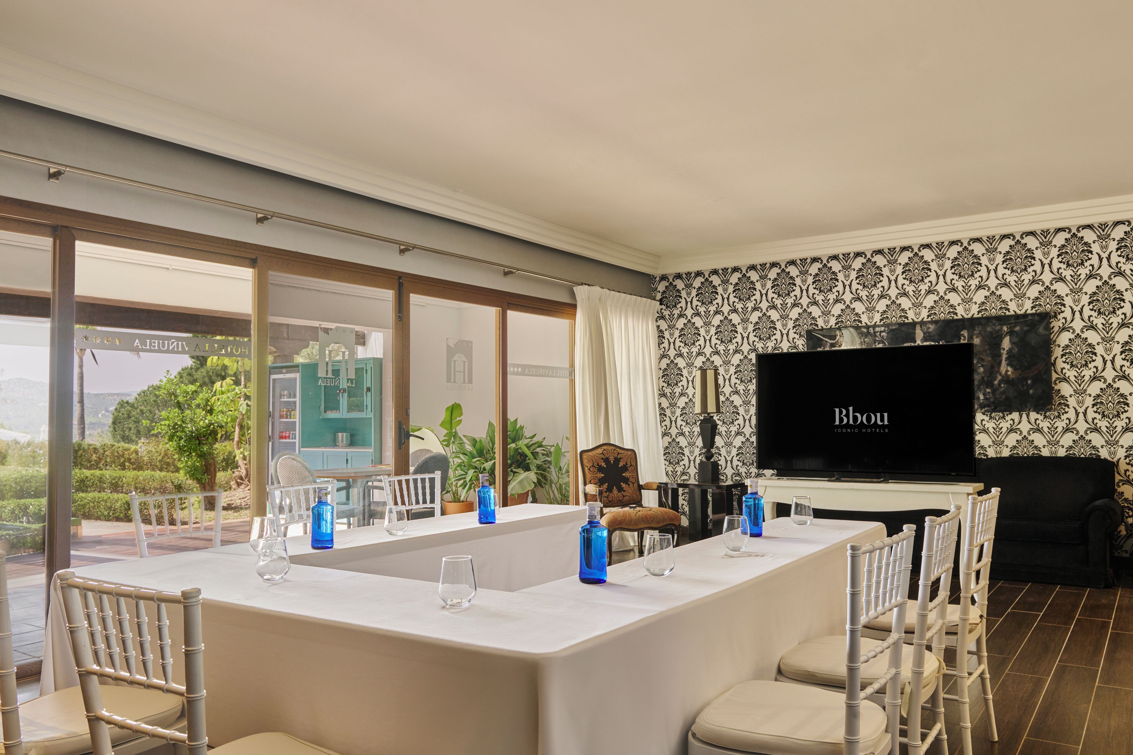 A modern hotel room with a large dining table, white chairs, a flat-screen TV, and a view of the outdoors.