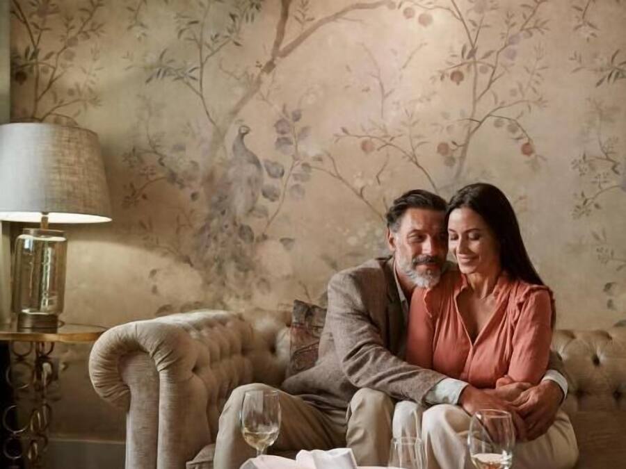 A couple sitting on a beige sofa in a room with floral wallpaper, a glass coffee table with wine glasses, and a lamp on a side table.
