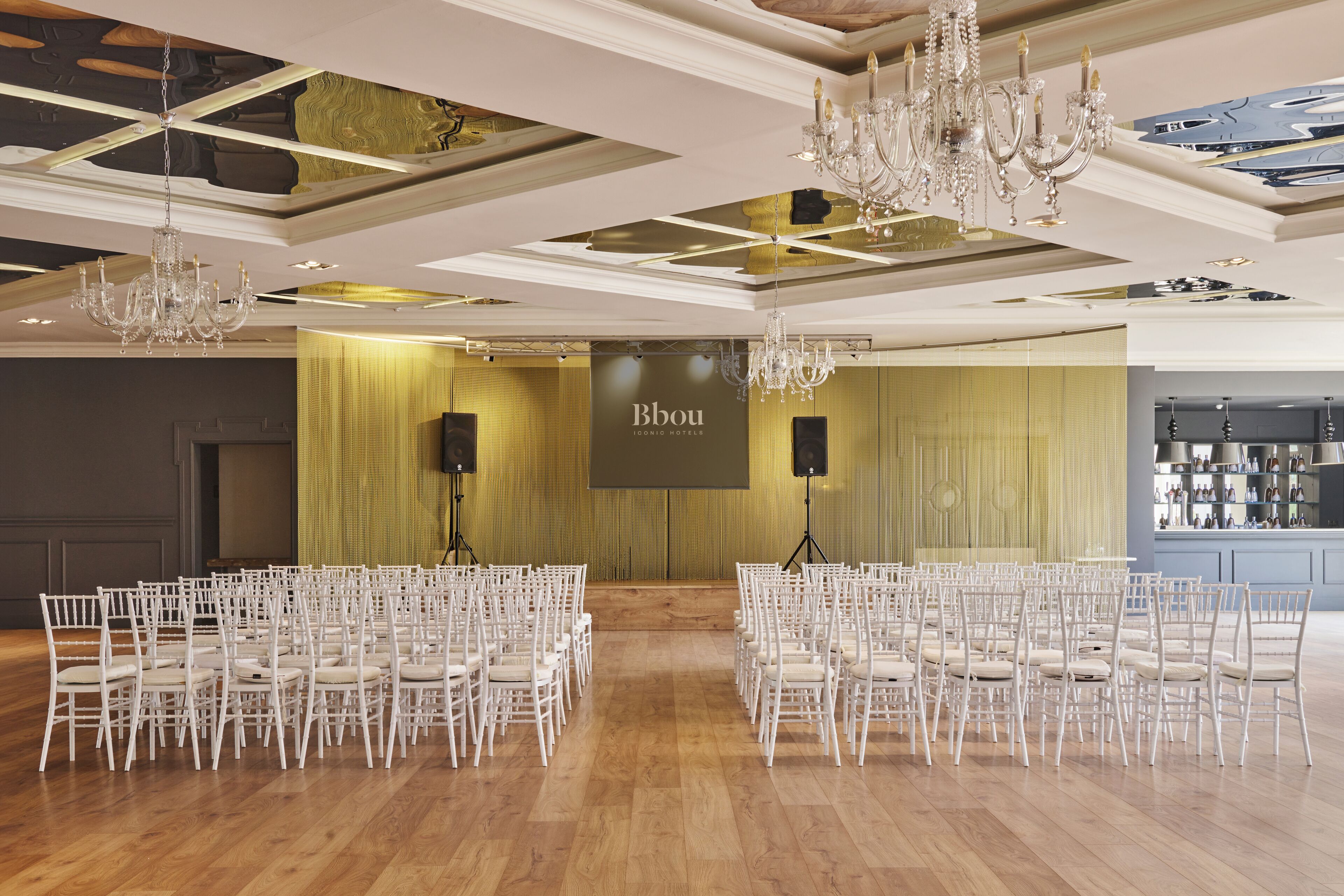 Un salón amplio con filas de sillas blancas dispuestas para un evento, una gran pantalla mostrando 'Bbou Iconic Hotel' y elegantes candelabros.