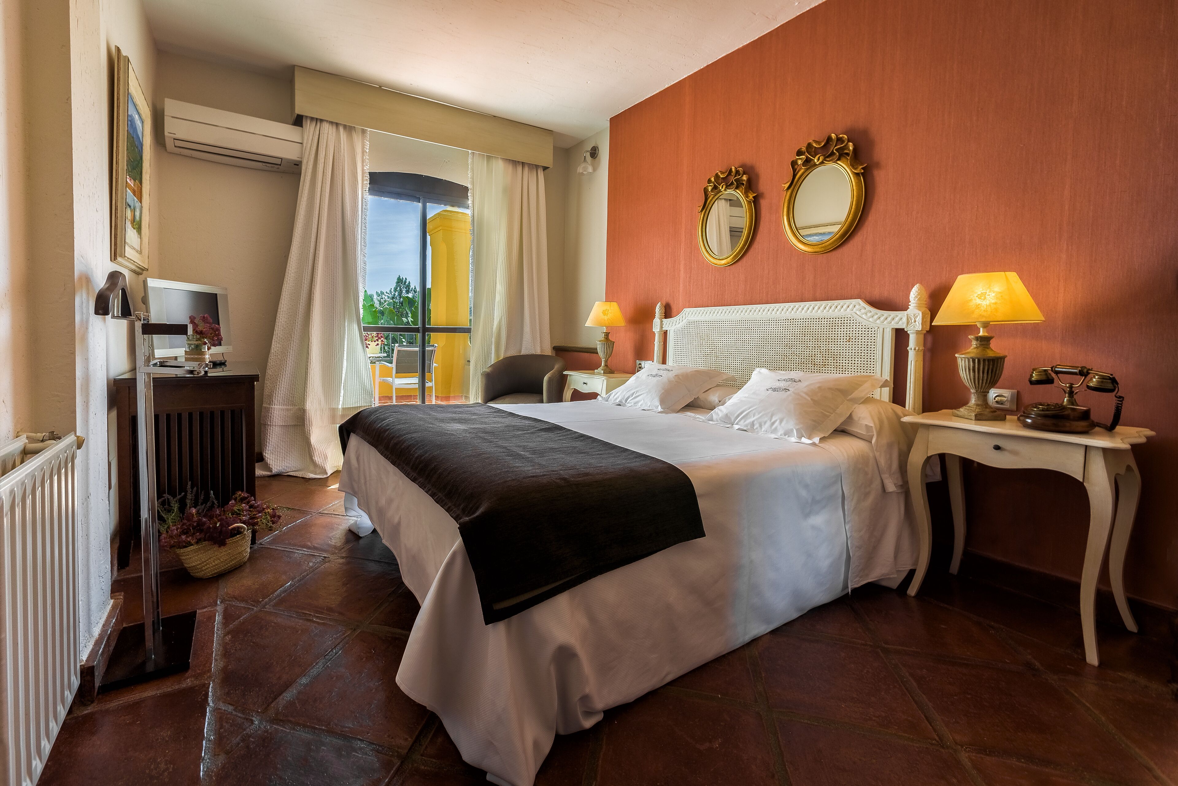 A hotel room with a large bed, two bedside lamps, a small table with a lamp, a chair, and a view of the outdoors.