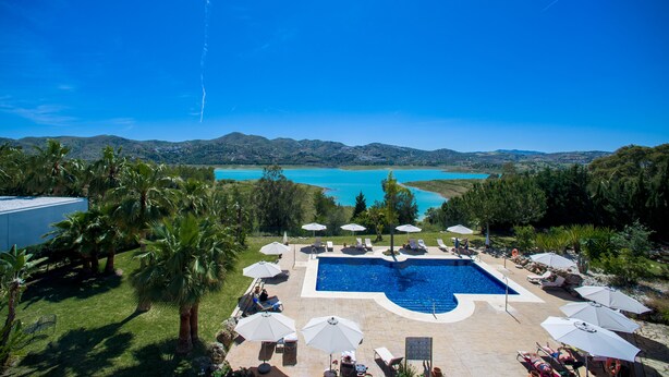En pool med udsigt over en sø og bjerge, omgivet af palmer og liggestole.