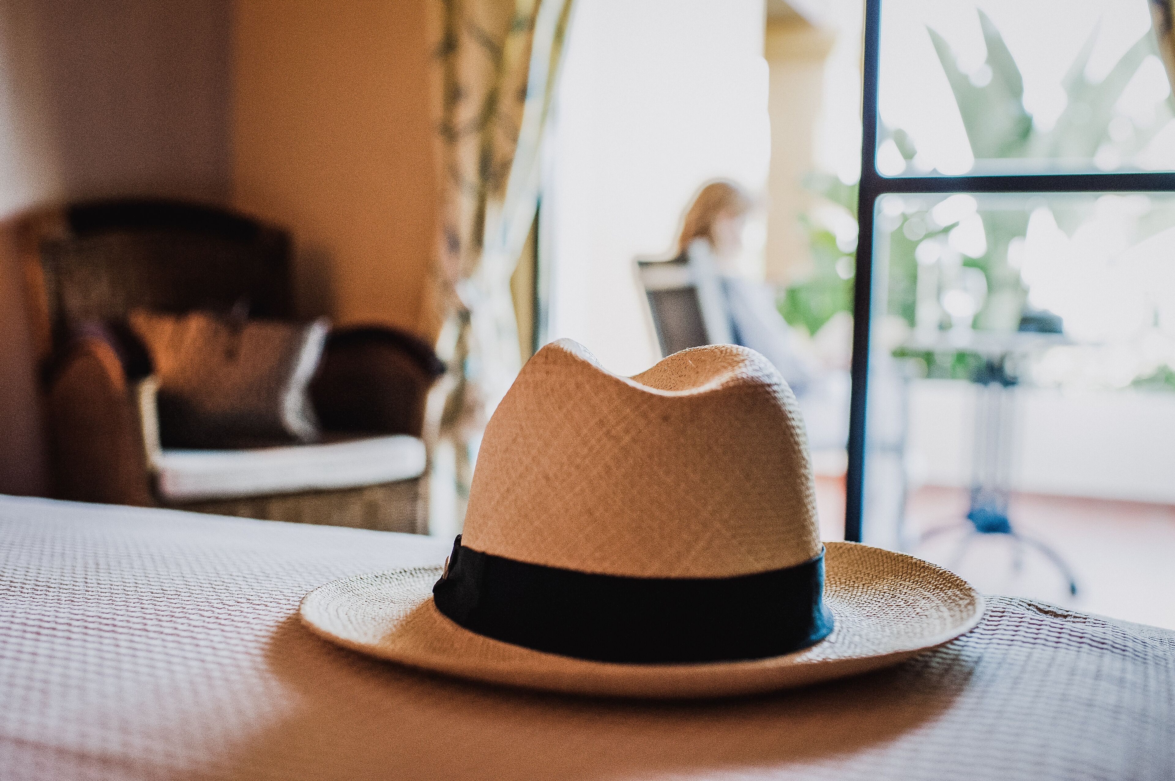 Un sombrero de paja con una banda negra sobre una mesa en una habitación con vistas a una persona sentada a otra mesa al fondo.