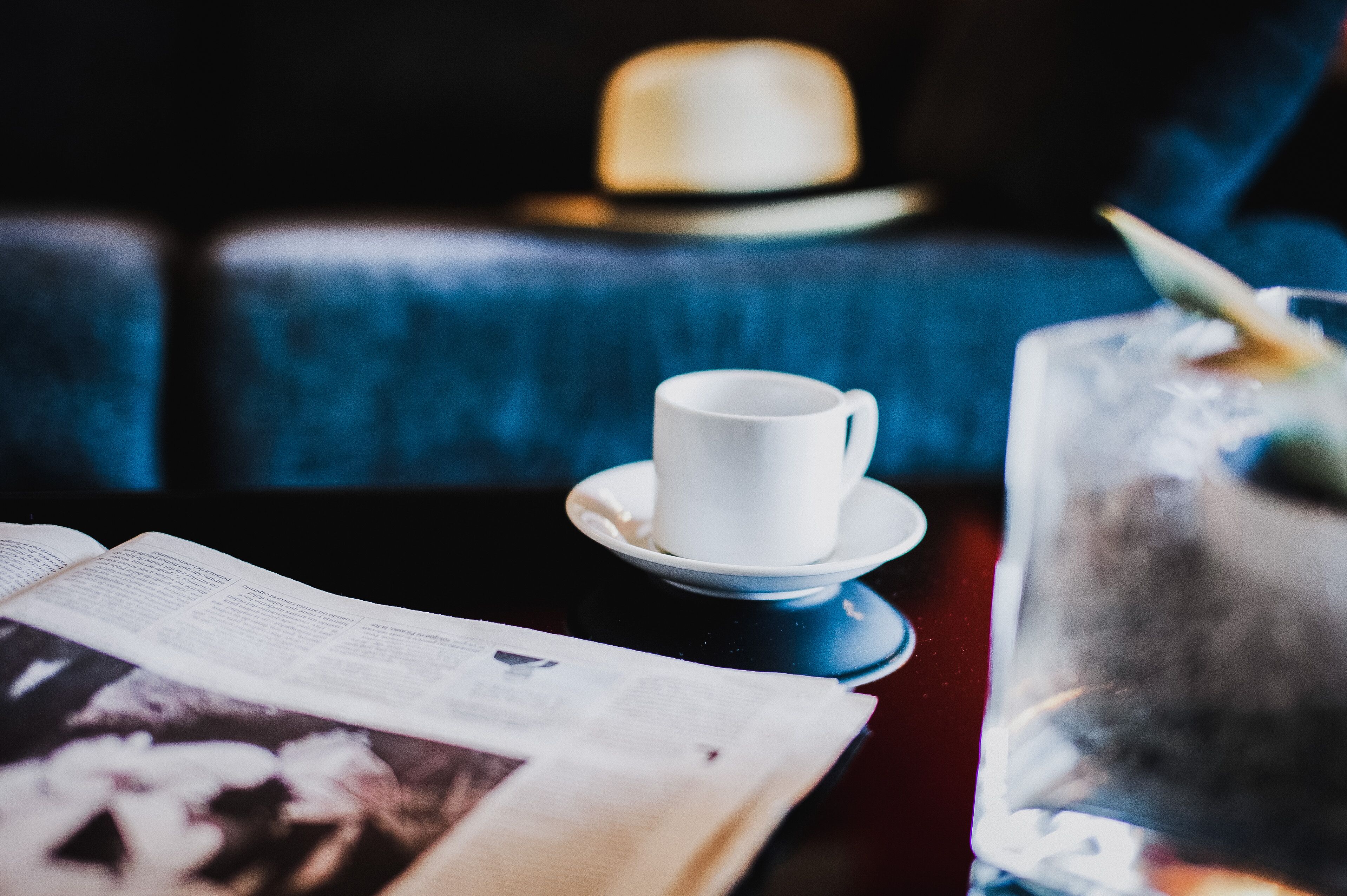A white cup on a saucer, a glass with a straw, and an open book on a table.