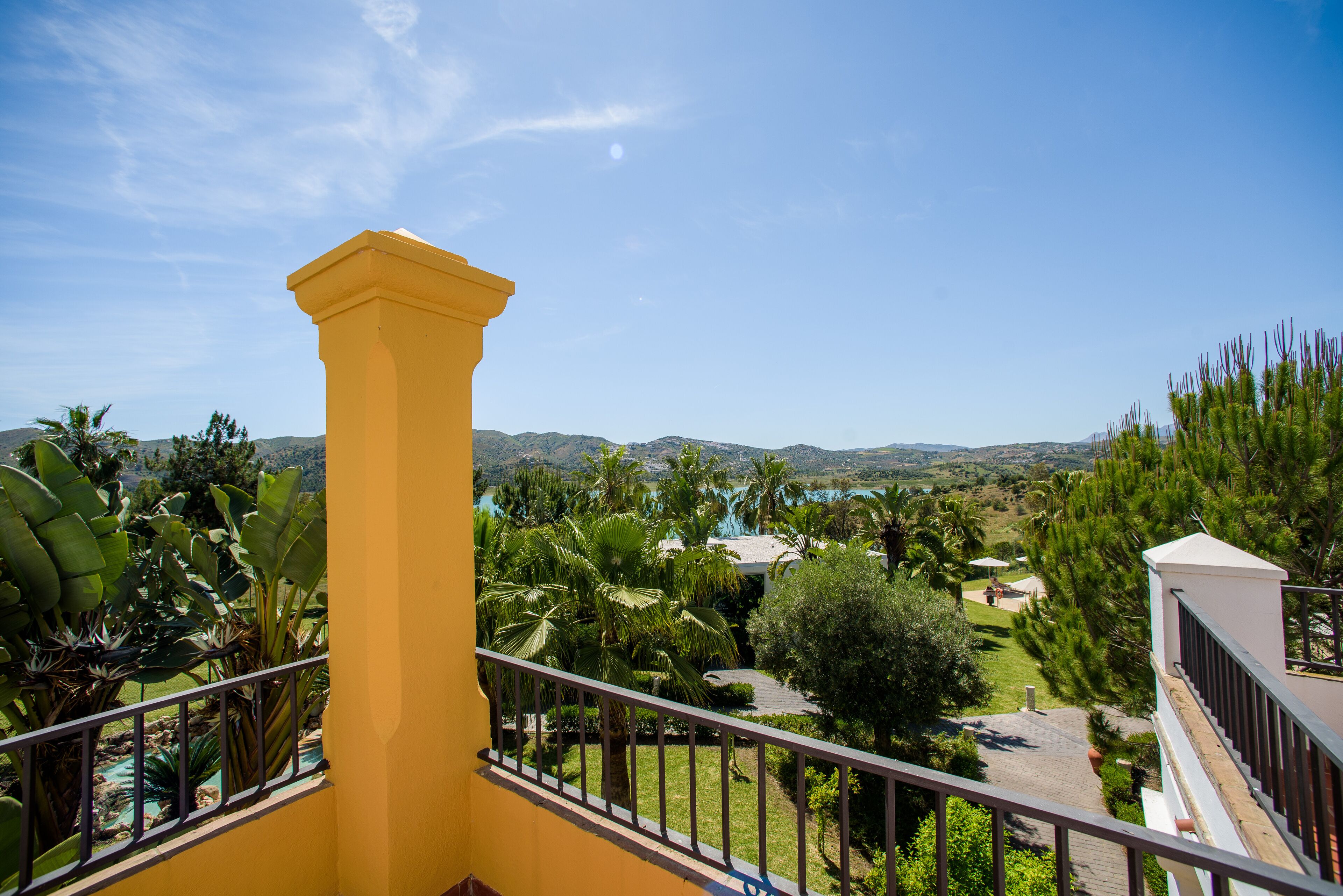 Un edificio amarillo con balcón que da a un área ajardinada con palmeras y vistas a colinas distantes.