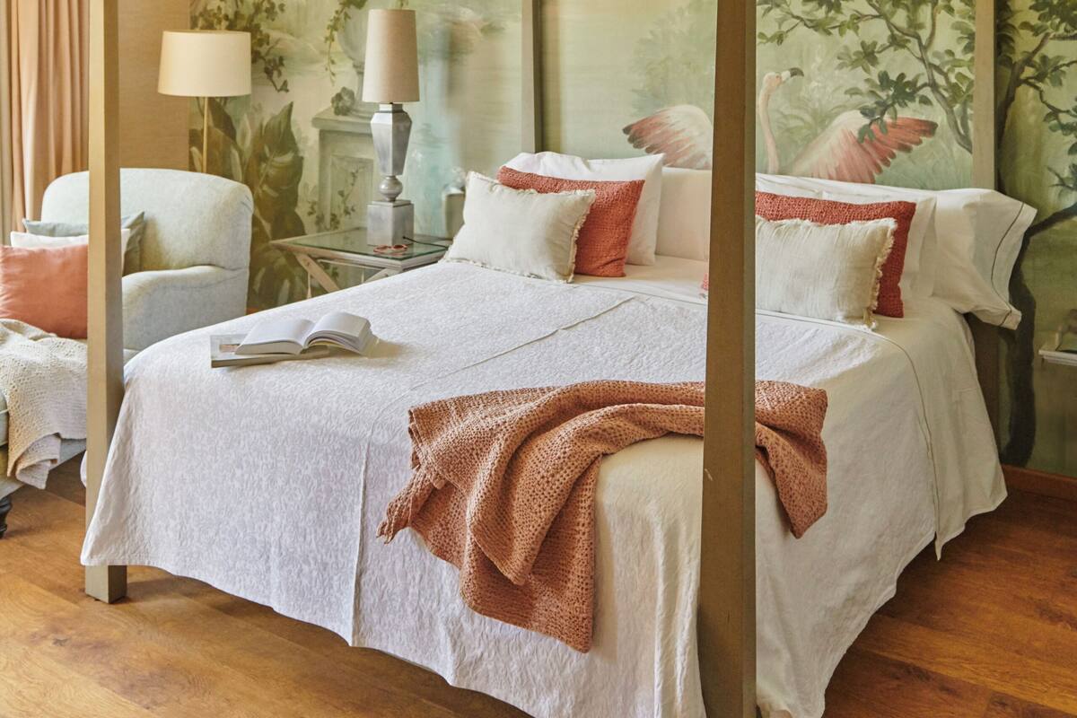 A four-poster bed with a white bedspread and red accents, a nightstand with a lamp, and a painting of a tropical scene on the wall.