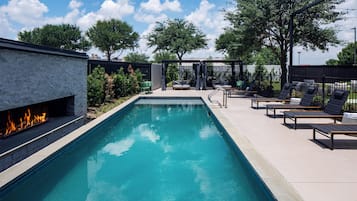 Piscina rectangular con una fuente de fuego en un lateral, rodeada de sillones de descanso y una zona de estar cubierta.