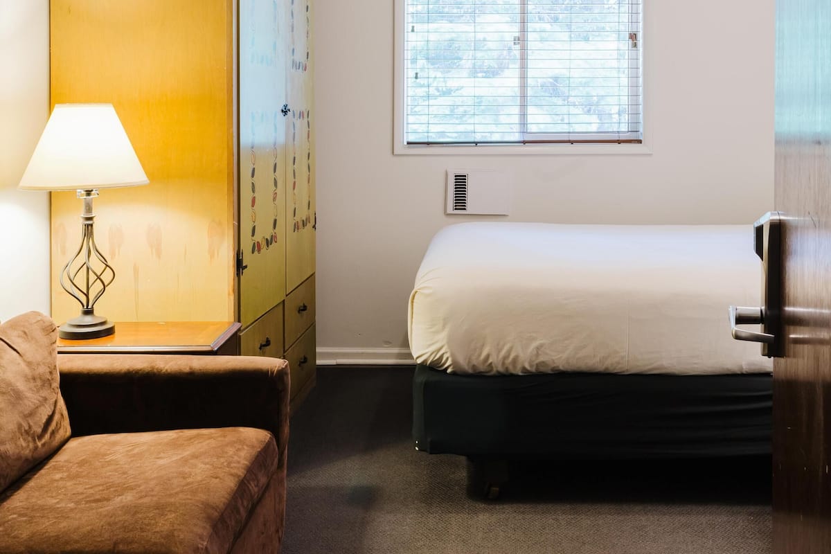 Una habitación de hotel con cama, sofá, lámpara, armario y ventana con persianas.