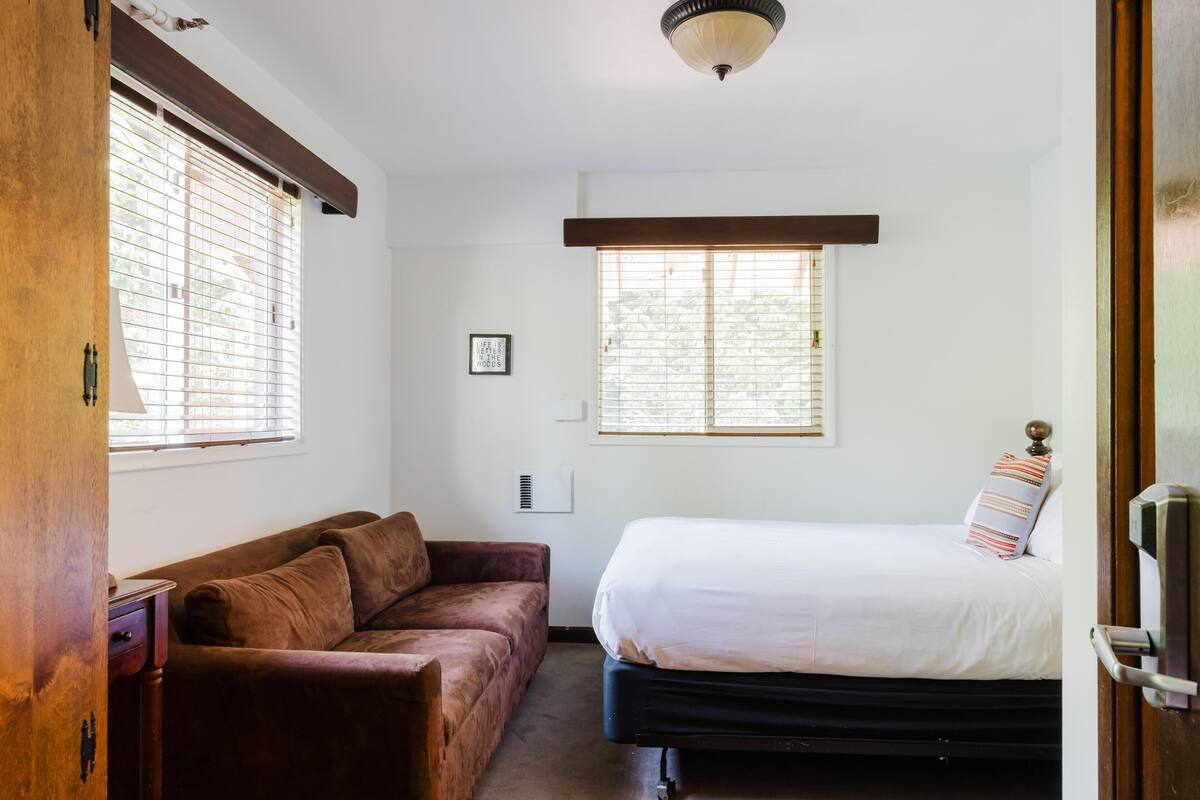 Un dormitorio con una cama, un sofá marrón y dos ventanas con persianas.