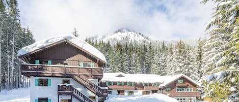 A chalet-style hotel with multiple levels, wooden balconies, and a snow-covered mountain backdrop.