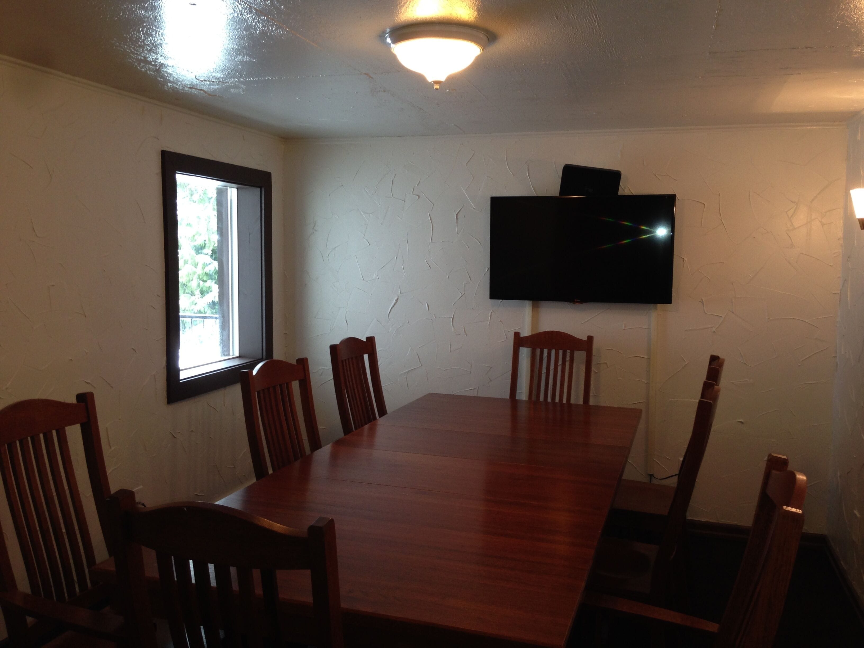 Un salón de conferencias con una mesa larga y seis sillas de madera, un televisor de pantalla plana en la pared y un ventanal que proporciona luz natural.