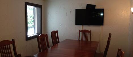 Un salón de conferencias con una mesa larga y seis sillas de madera, un televisor de pantalla plana en la pared y un ventanal que proporciona luz natural.