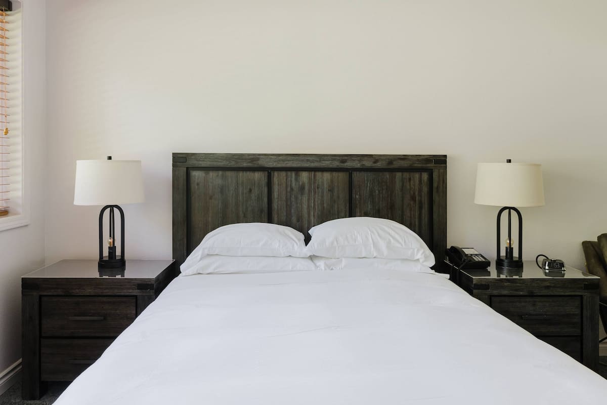 Una cama bien hecha con sábanas blancas, dos mesitas de noche con lámparas, un cabecero de madera y un teléfono sobre la mesa de la derecha.