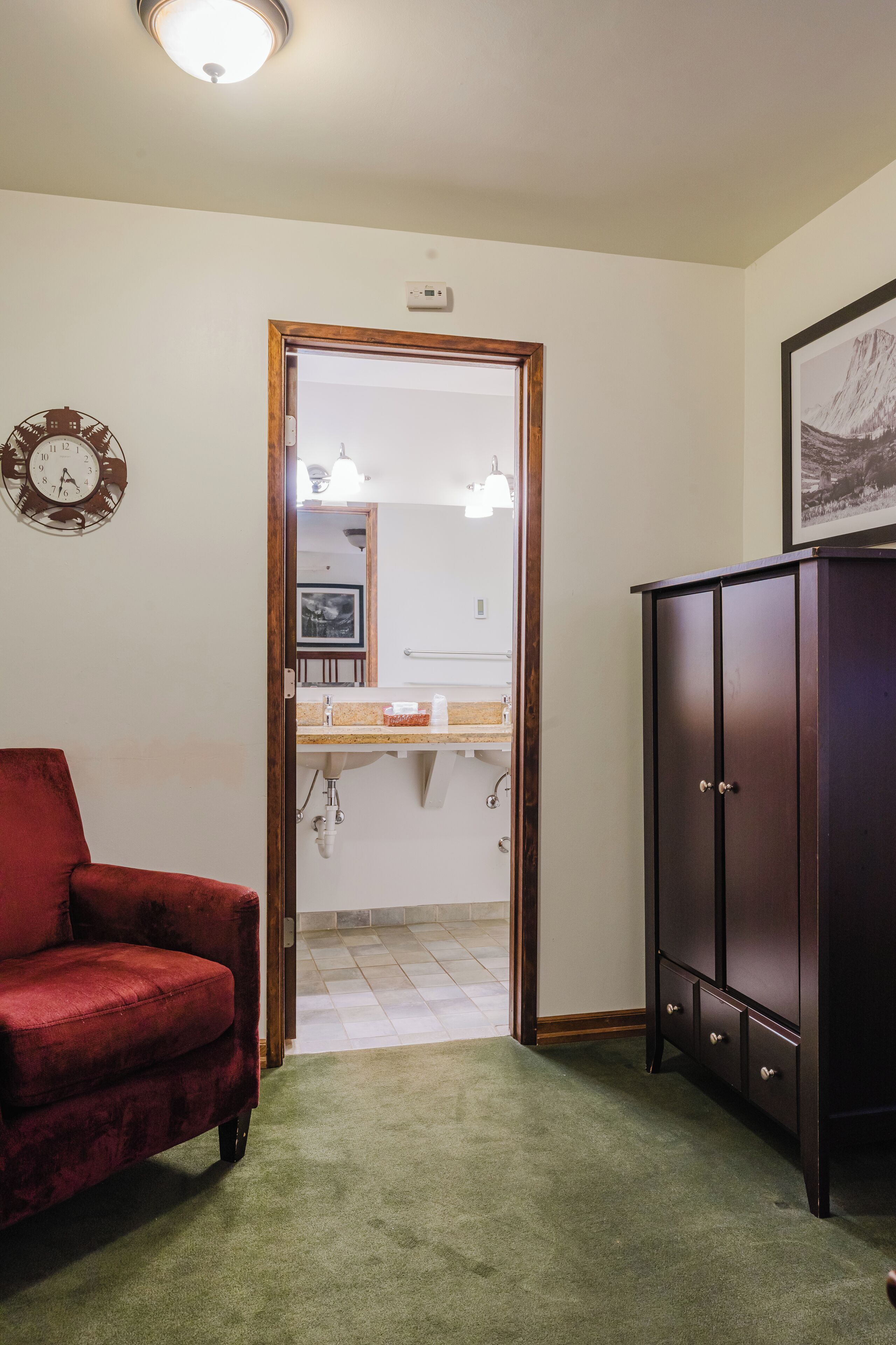 A room with a red chair, a wooden cabinet, and a doorway leading to a bathroom with a sink and mirror.