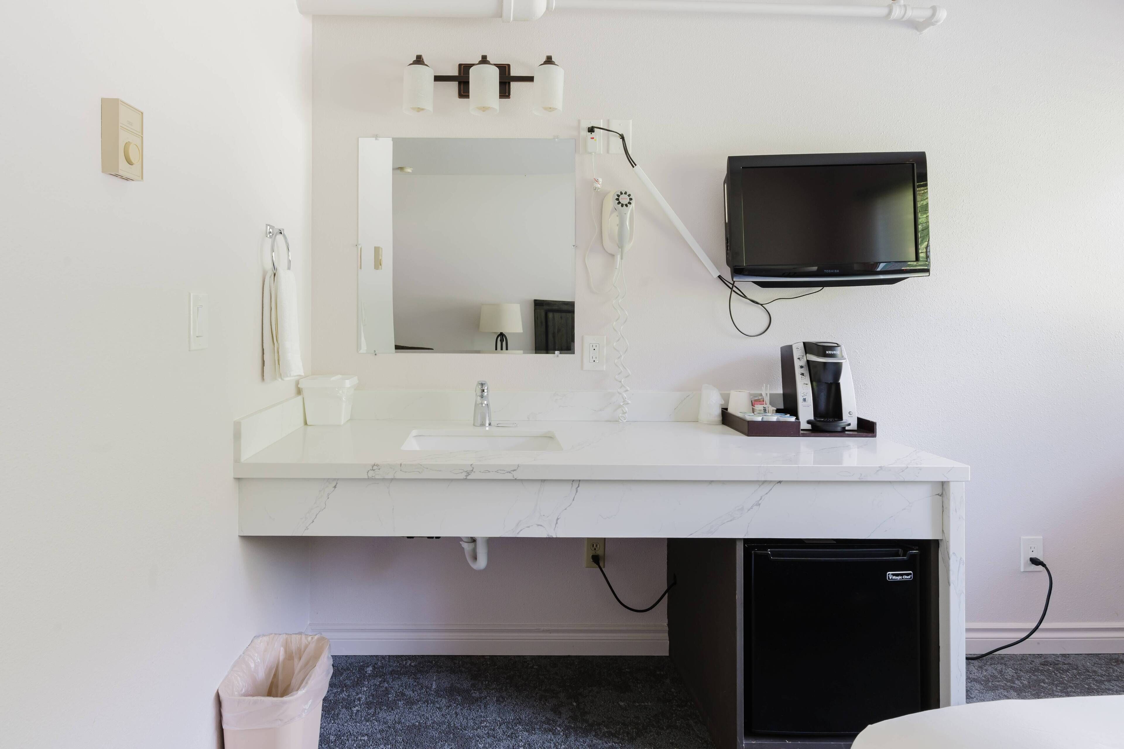A modern bathroom with a white marble sink, a mirror, a hairdryer, a TV mounted on the wall, and a small black refrigerator.