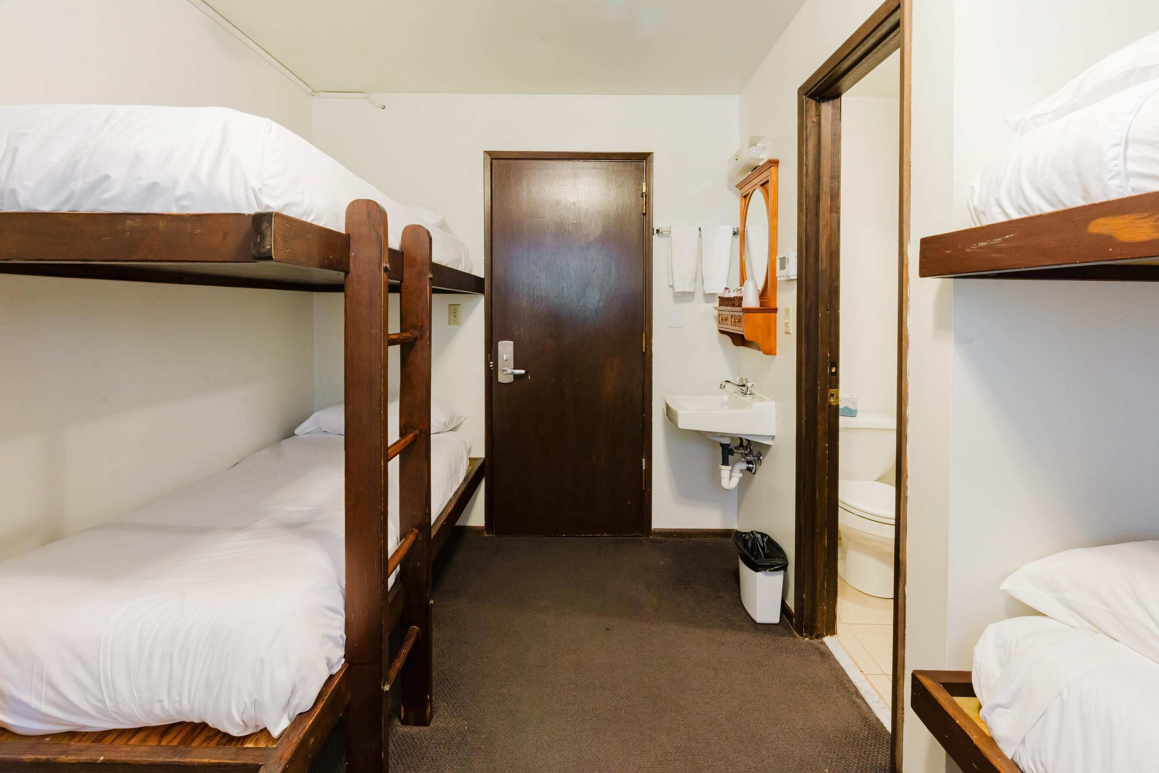 Un couloir étroit, avec des lits superposés de chaque côté, une porte au fond et un lavabo avec un miroir et un porte-serviettes sur le mur de droite.