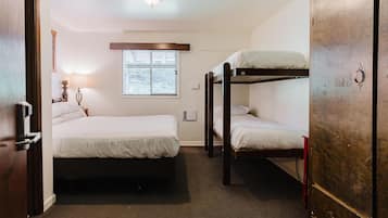 Quarto com uma cama de solteiro, uma cama beliche, uma janela com persianas, uma luminária e uma porta.