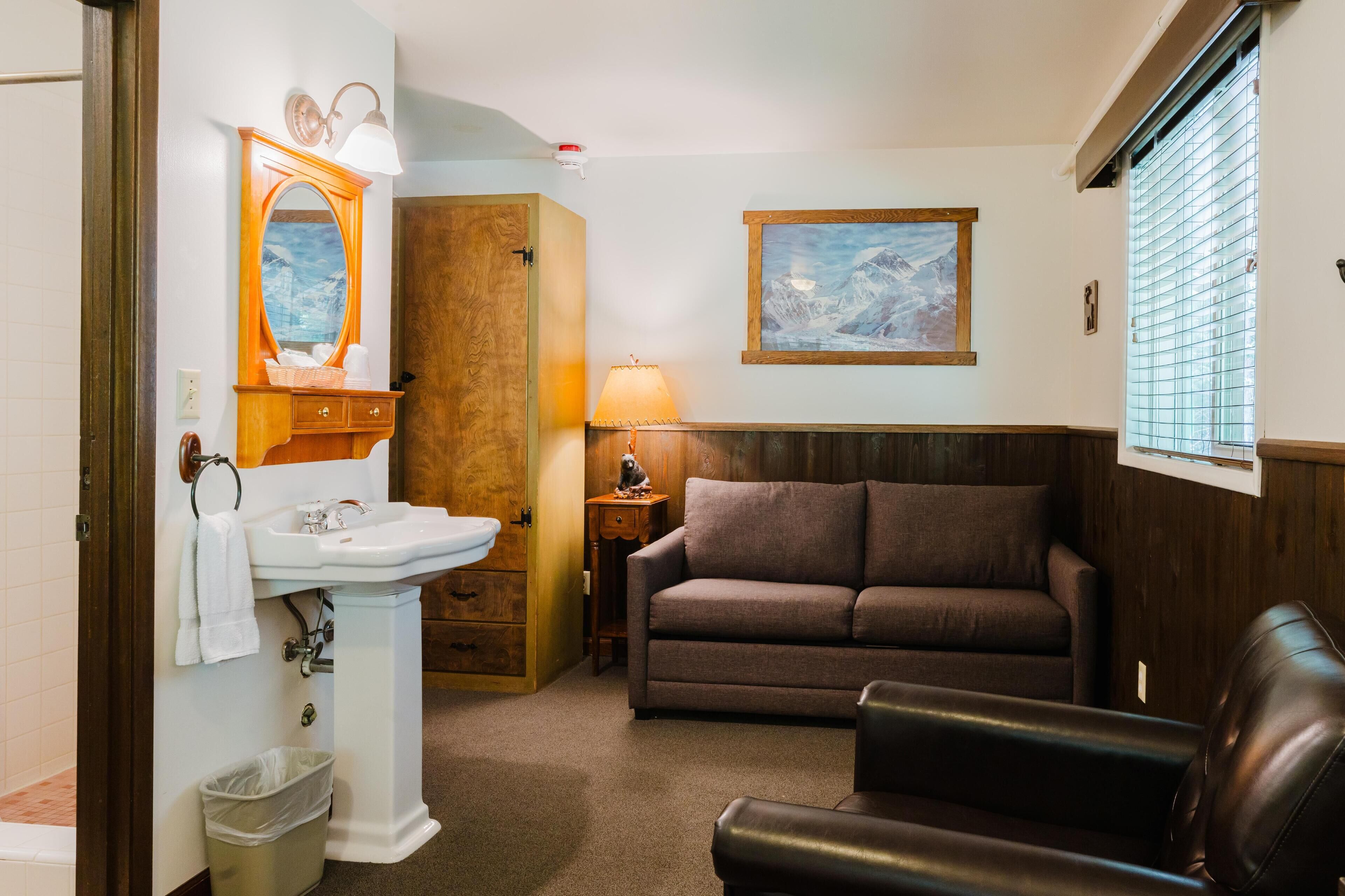 A bathroom with a sink, mirror, and wooden cabinet. There is a brown couch and a black leather chair.