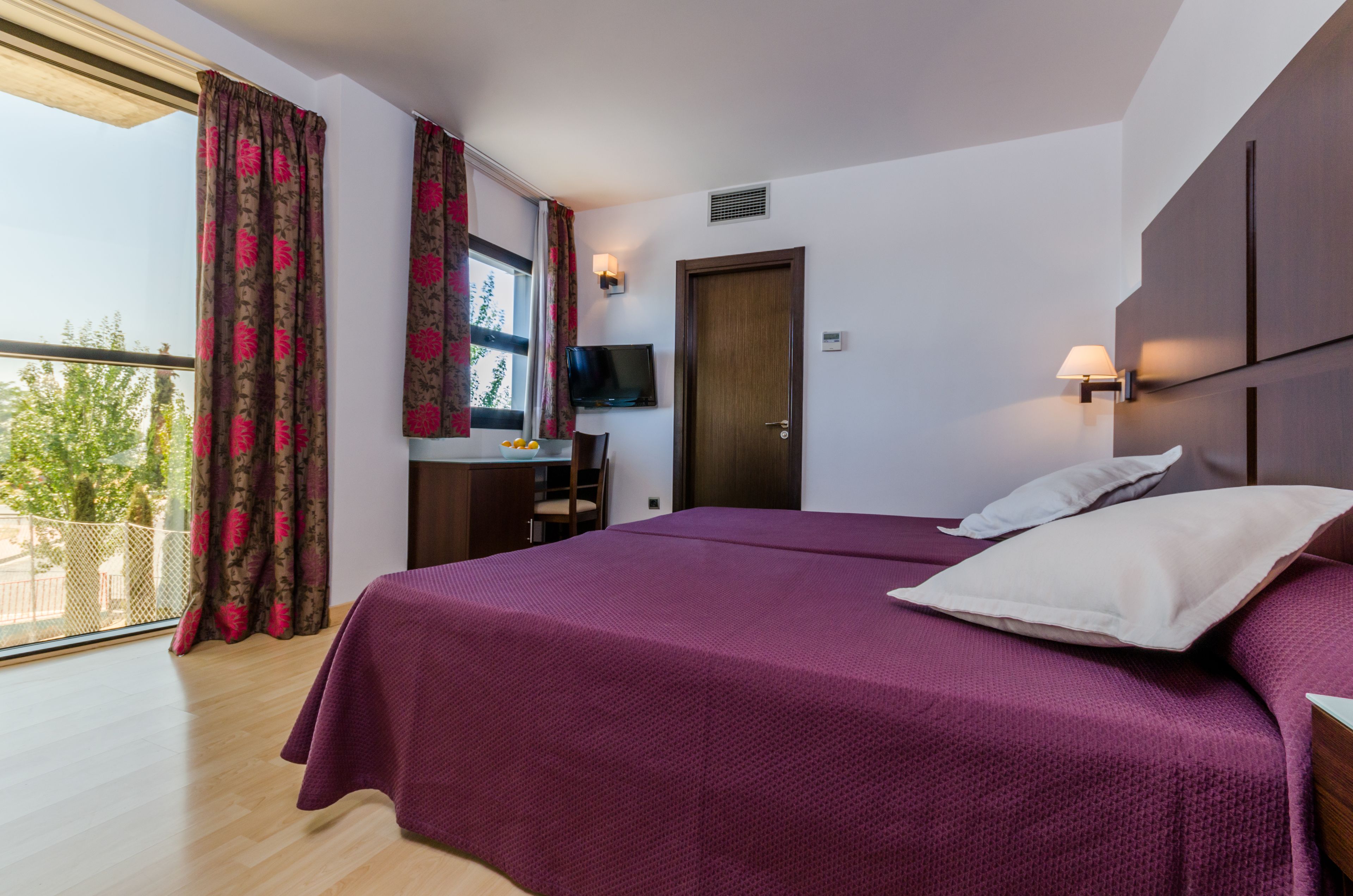 Habitación de hotel con una cama grande, un escritorio con silla, televisión y un balcón con cortinas.
