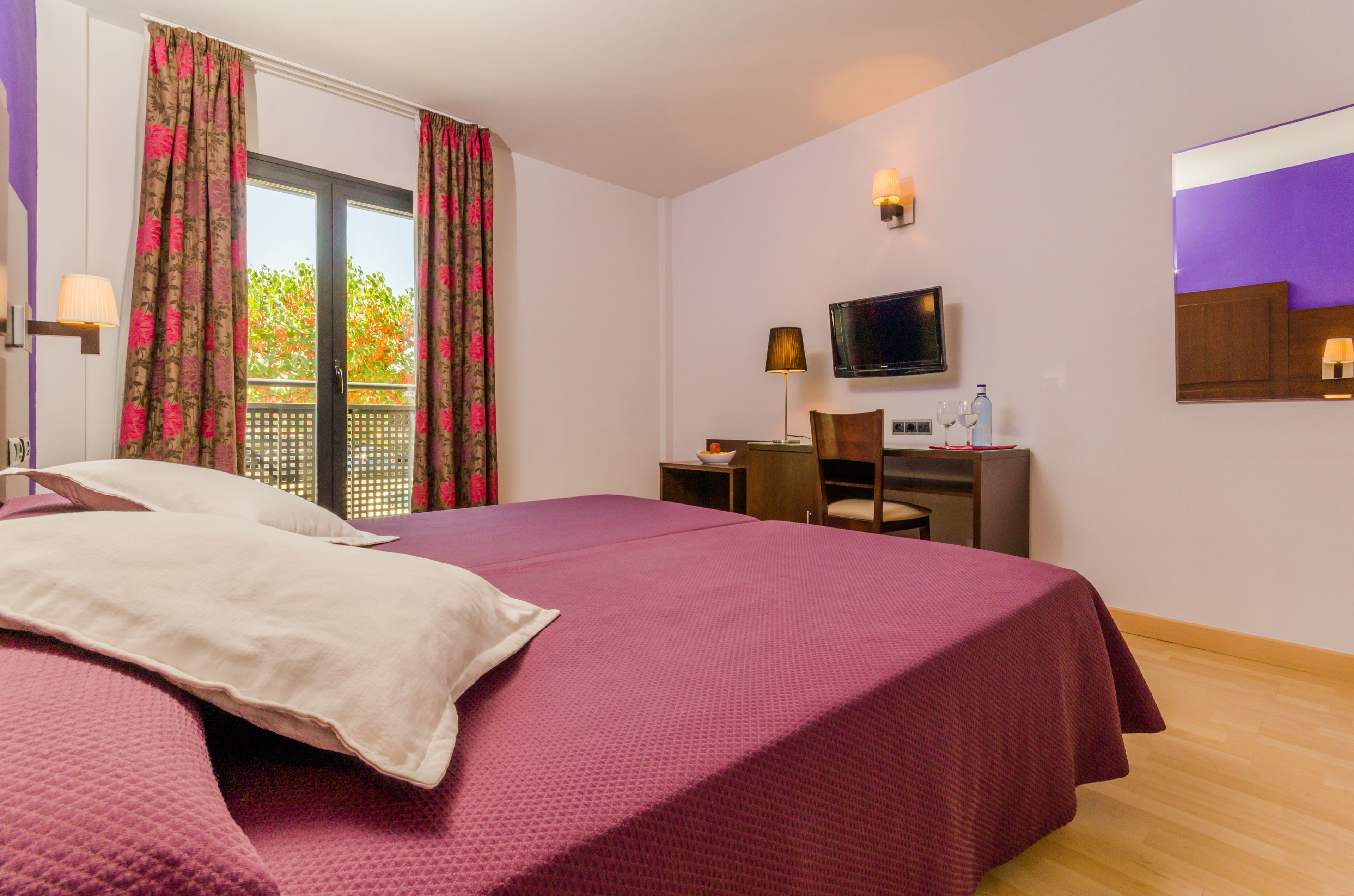 A hotel room with a large bed, a desk with a chair, a television, and a window with curtains.