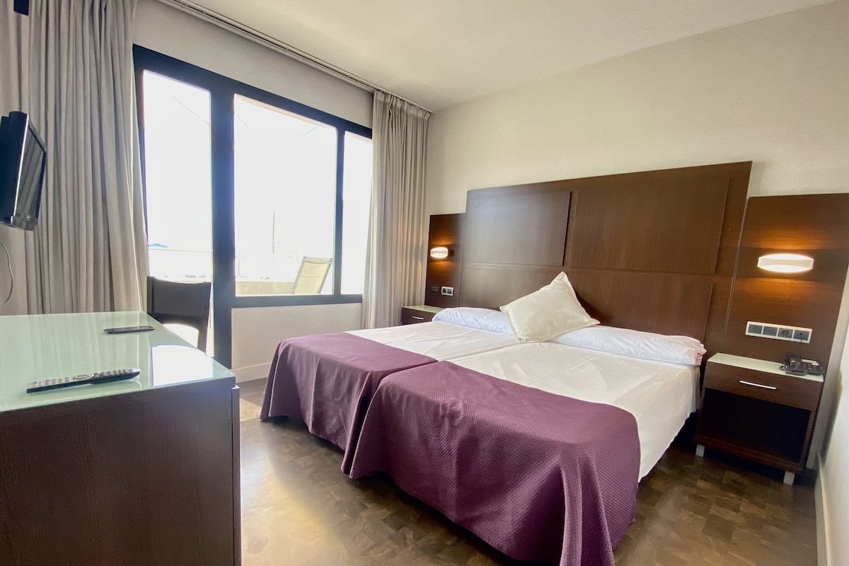 Quarto de hotel com uma cama grande, uma escrivaninha com cadeira, uma janela com cortinas e uma mesa de cabeceira com abajur.