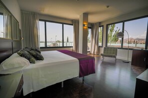Une chambre d’hôtel équipée d’un lit, d’une chaise, d’un bureau et de grandes fenêtres donnant sur un plan d’eau et sur des collines lointaines.