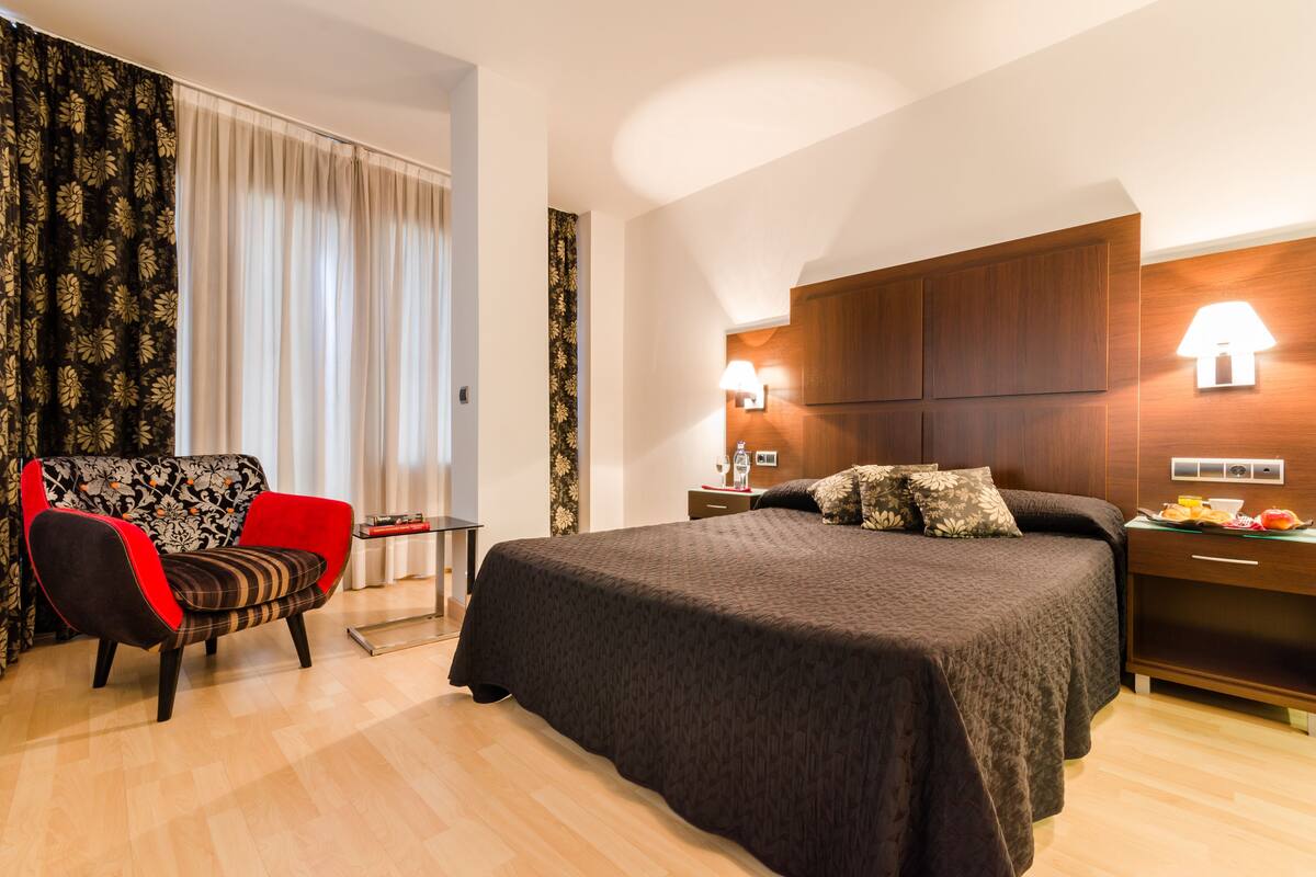 Quarto de hotel com uma cama grande, uma poltrona preta e vermelha, uma mesa de cabeceira com abajur e vista para o exterior através de cortinas leves.