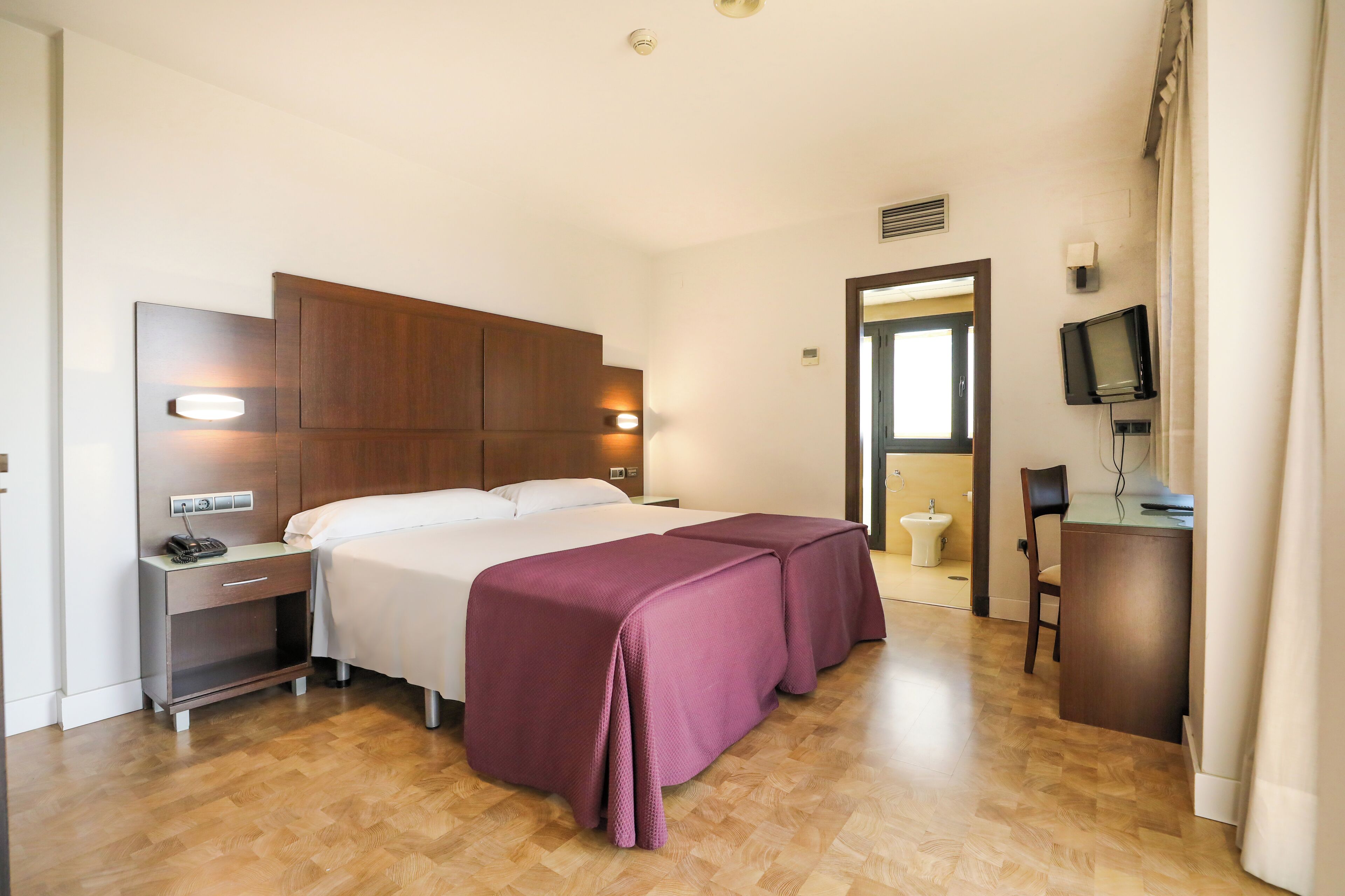 A hotel room with a bed, bedside table, TV, and a desk.