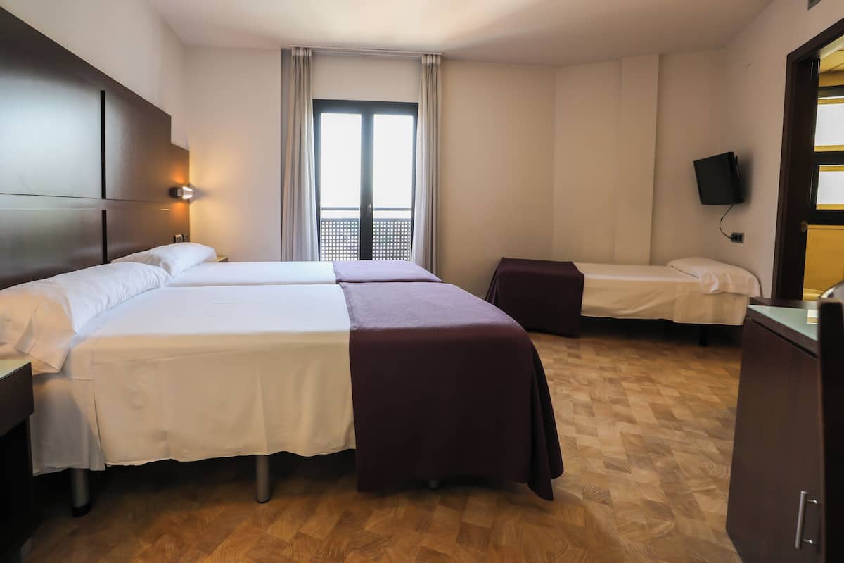 Quarto de hotel com três camas, piso de madeira, uma janela grande com cortinas, televisão de tela plana na parede e uma escrivaninha com cadeira.