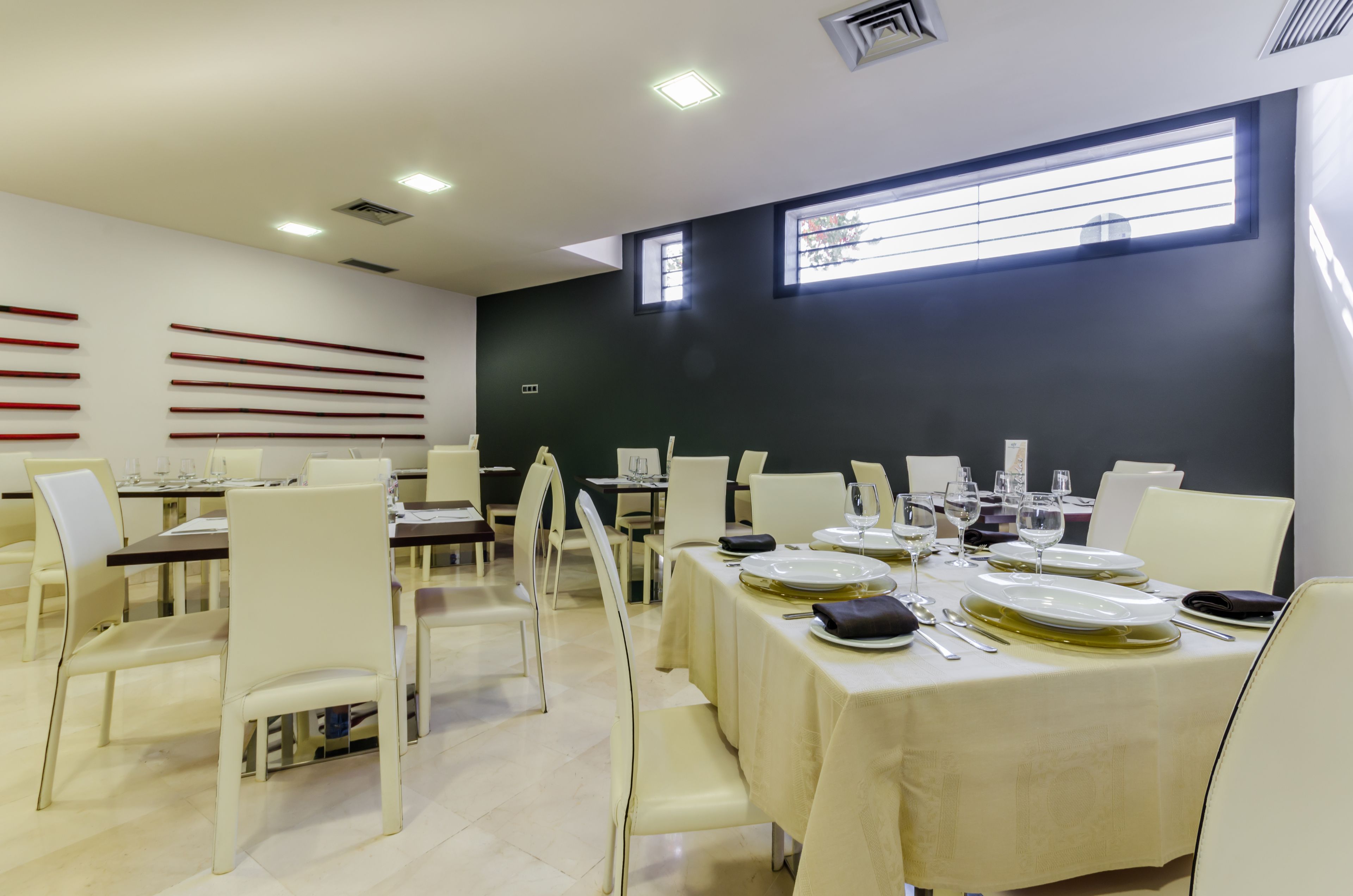 Zona pranzo con tavoli apparecchiati per quattro persone, sedie bianche e una parete nera con una finestra.