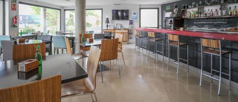 A modern bar area with a red countertop, bar stools, and a variety of bottles and glasses. There are tables and chairs arranged for dining, and a television mounted on the wall.