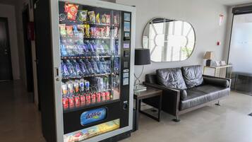 A vending machine with snacks and drinks, a sofa, a lamp, and a wall-mounted mirror in a hotel lobby.