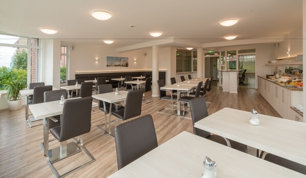 Un espace repas moderne doté de plusieurs tables et chaises, d’un coin cuisine et de grandes fenêtres offrant une vue sur la verdure.