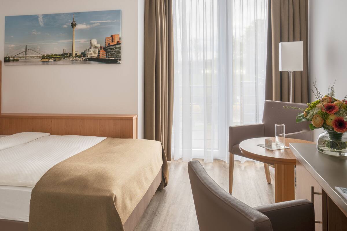 A hotel room with a bed, a desk with a chair, a window with curtains, and a painting of a cityscape on the wall.