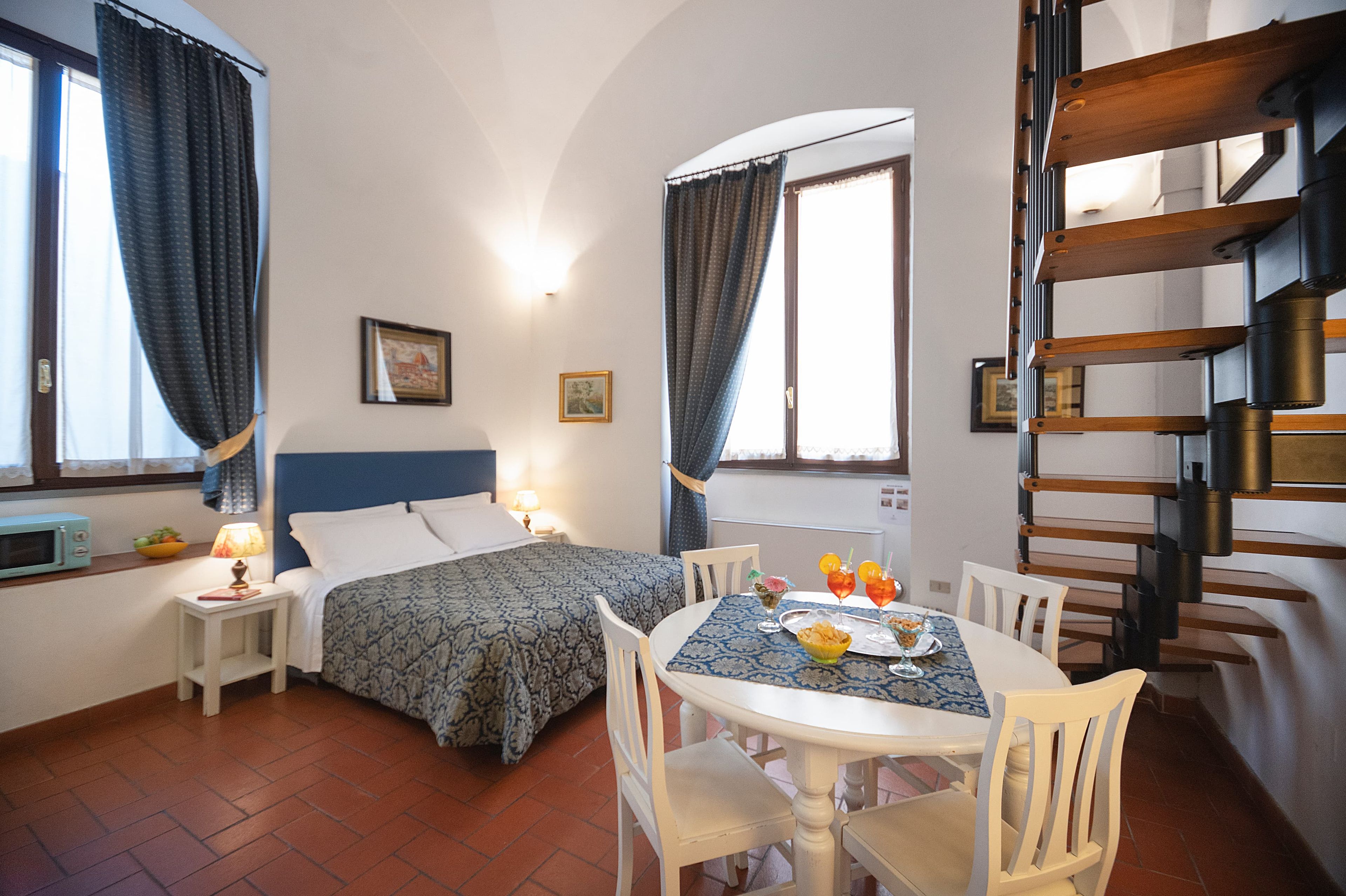 A hotel room with a bed, a dining table with chairs, a small kitchen area with a microwave, and a staircase.