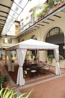 A covered outdoor seating area with a table and chairs, surrounded by potted plants and a white canopy.