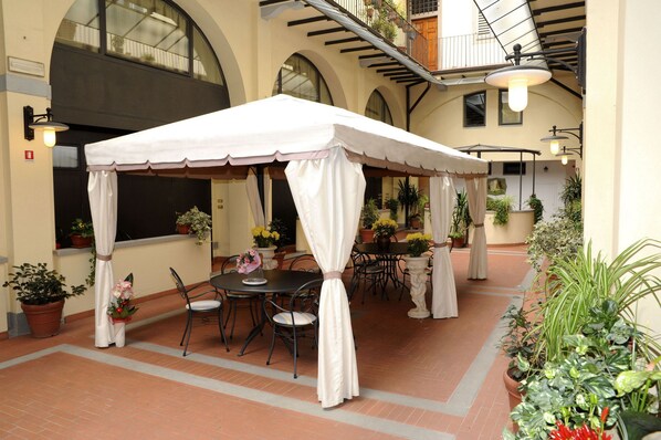 A covered outdoor dining area with tables and chairs, surrounded by potted plants and a balcony with railings.