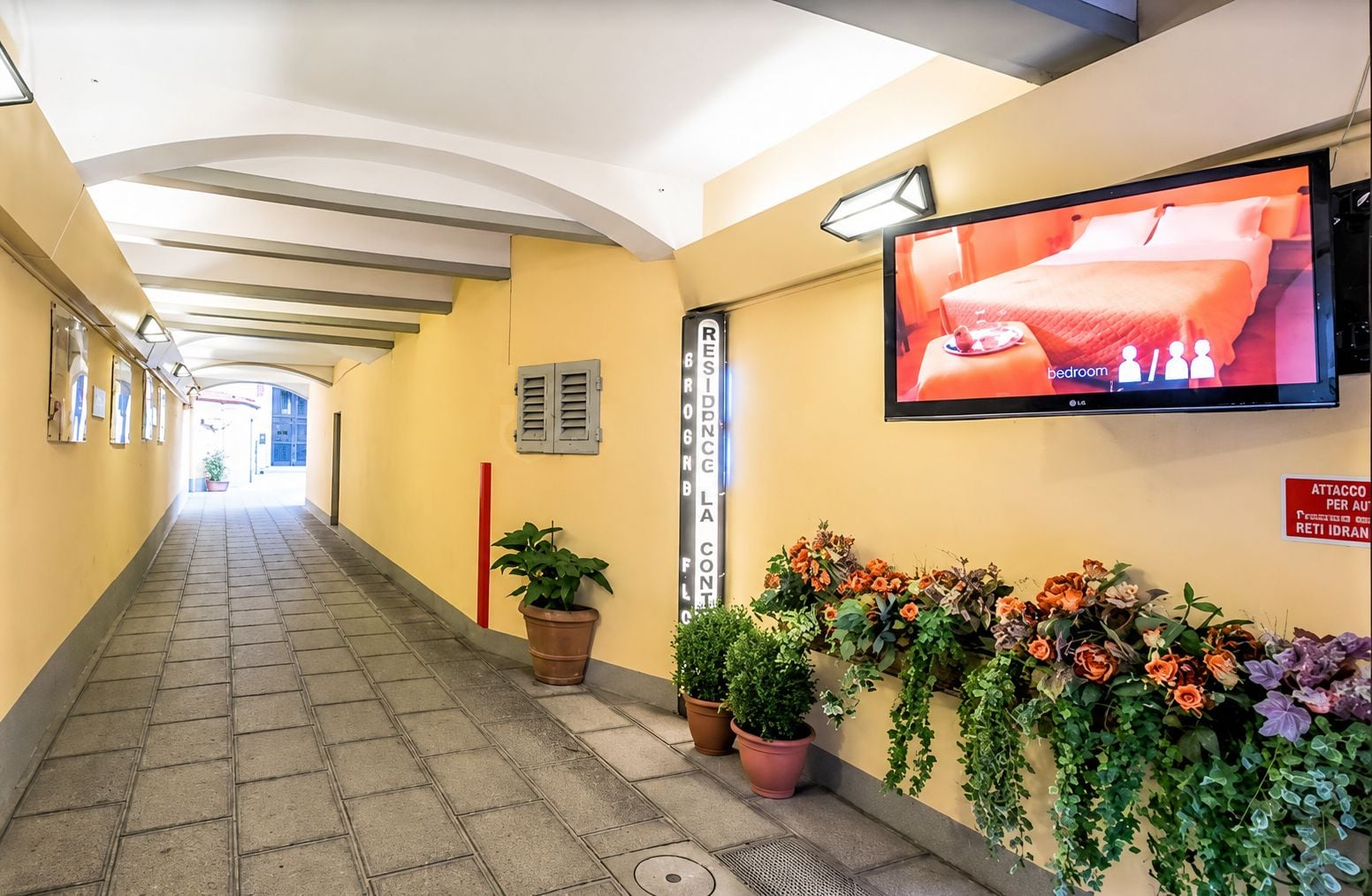 Pasillo de hotel con un televisor mostrando un dormitorio, plantas en macetas y un cartel que indica una residencia para personas mayores.