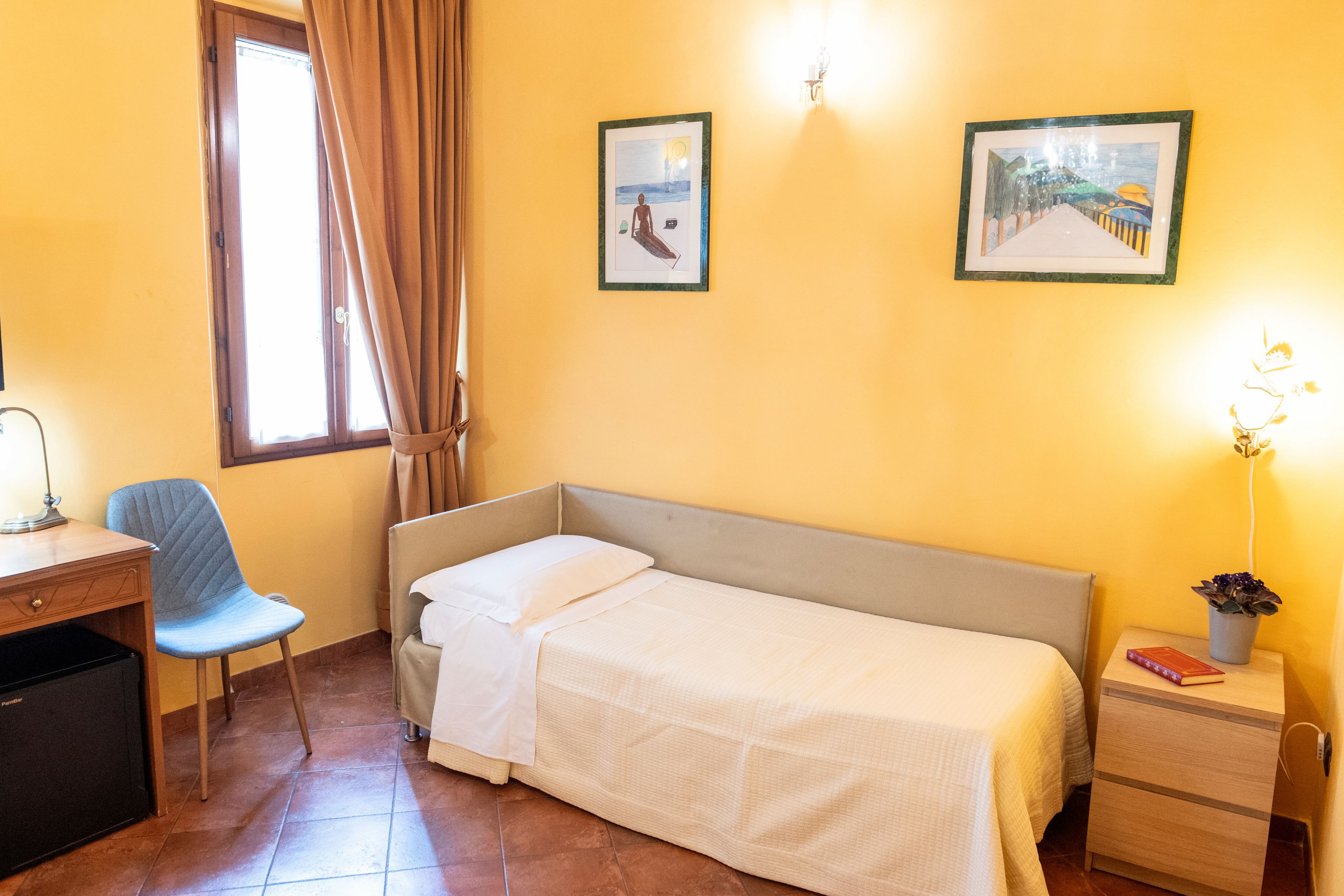 A single bed with a headboard, a desk with a chair, a small bedside table with a lamp, and two framed pictures on the wall.