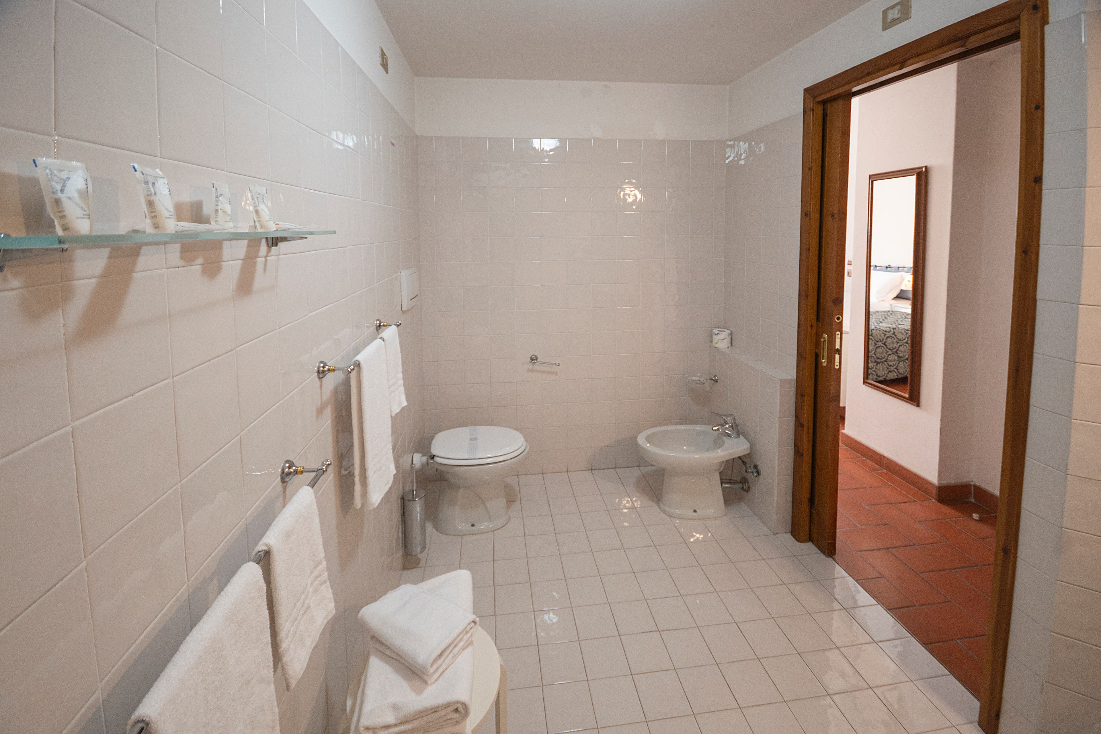 Un baño con paredes de azulejos blancos, un inodoro, un bidé y un estante de vidrio con artículos de aseo.