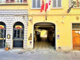 Un vicolo stretto con un edificio giallo a destra, una lavagna con scritte e un segnale che indica un parcheggio.