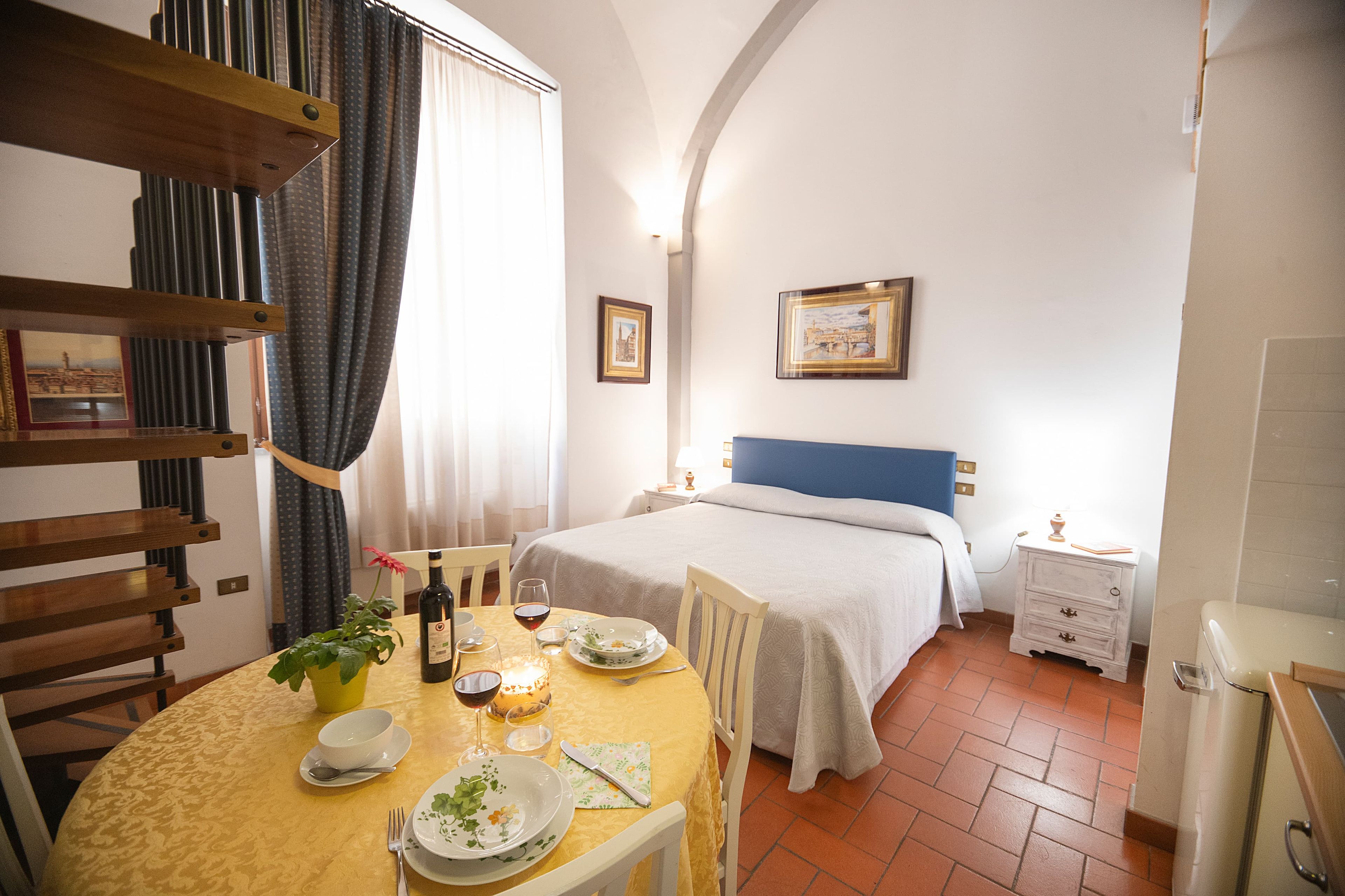 A hotel room with a bed, a dining table set for two, a chair, a bedside table with a lamp, and a wall-mounted picture.